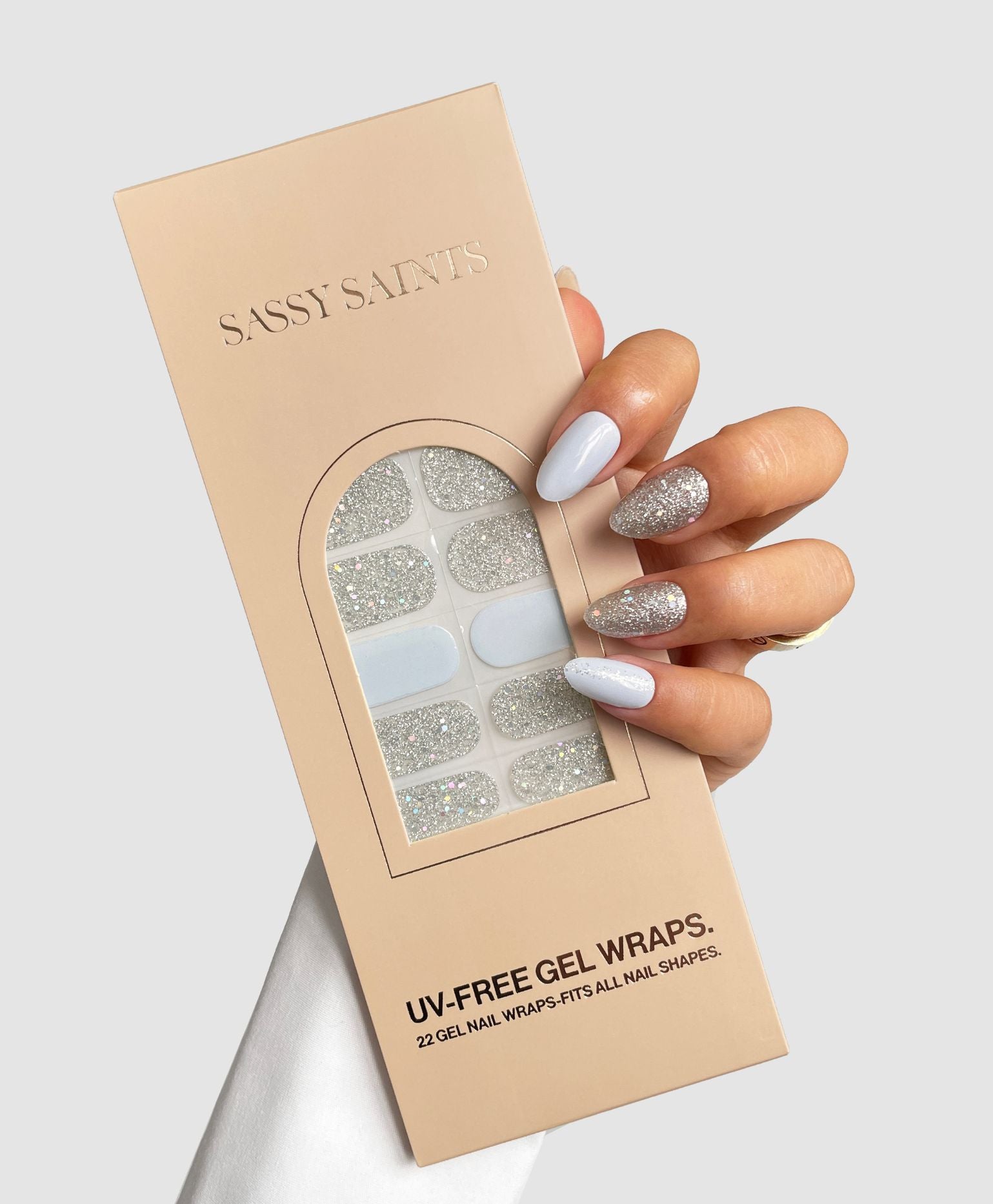 A hand with silver glitter and pale lavender nails holds a beige Sassy Saints - Brings The Salon Home package labeled Hazy Frost Gel Wrap, featuring UV-free gel nail wraps in silver glitter and pale colors.