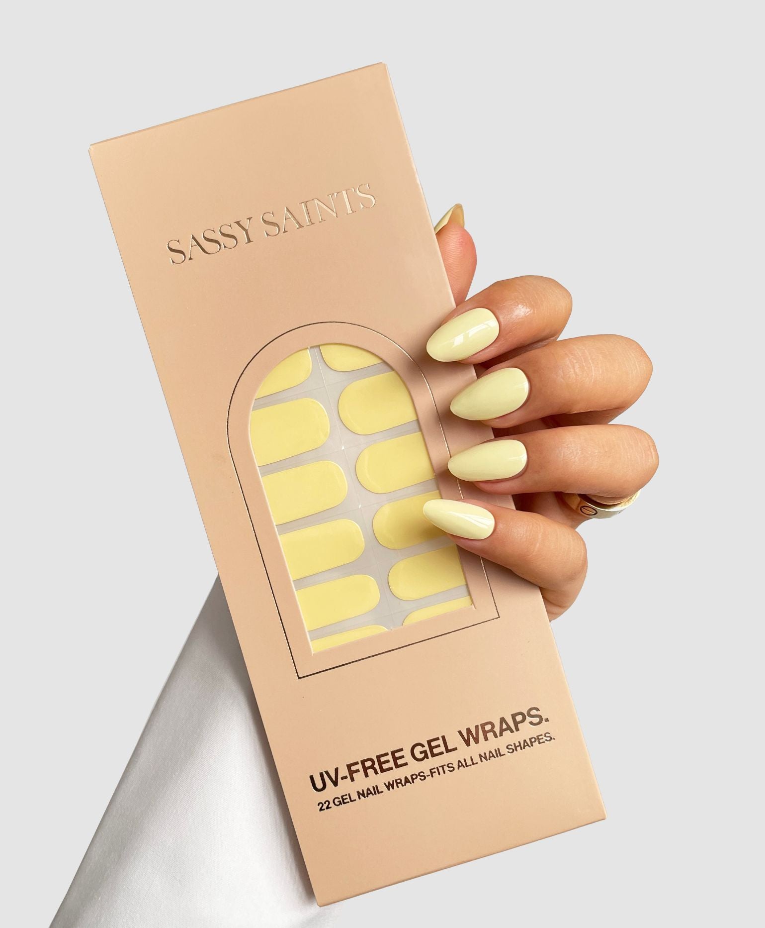 A hand with pale yellow manicured nails holds a Sassy Saints - Brings The Salon Home Buttercup Yellow Gel Wrap package against a plain light background.