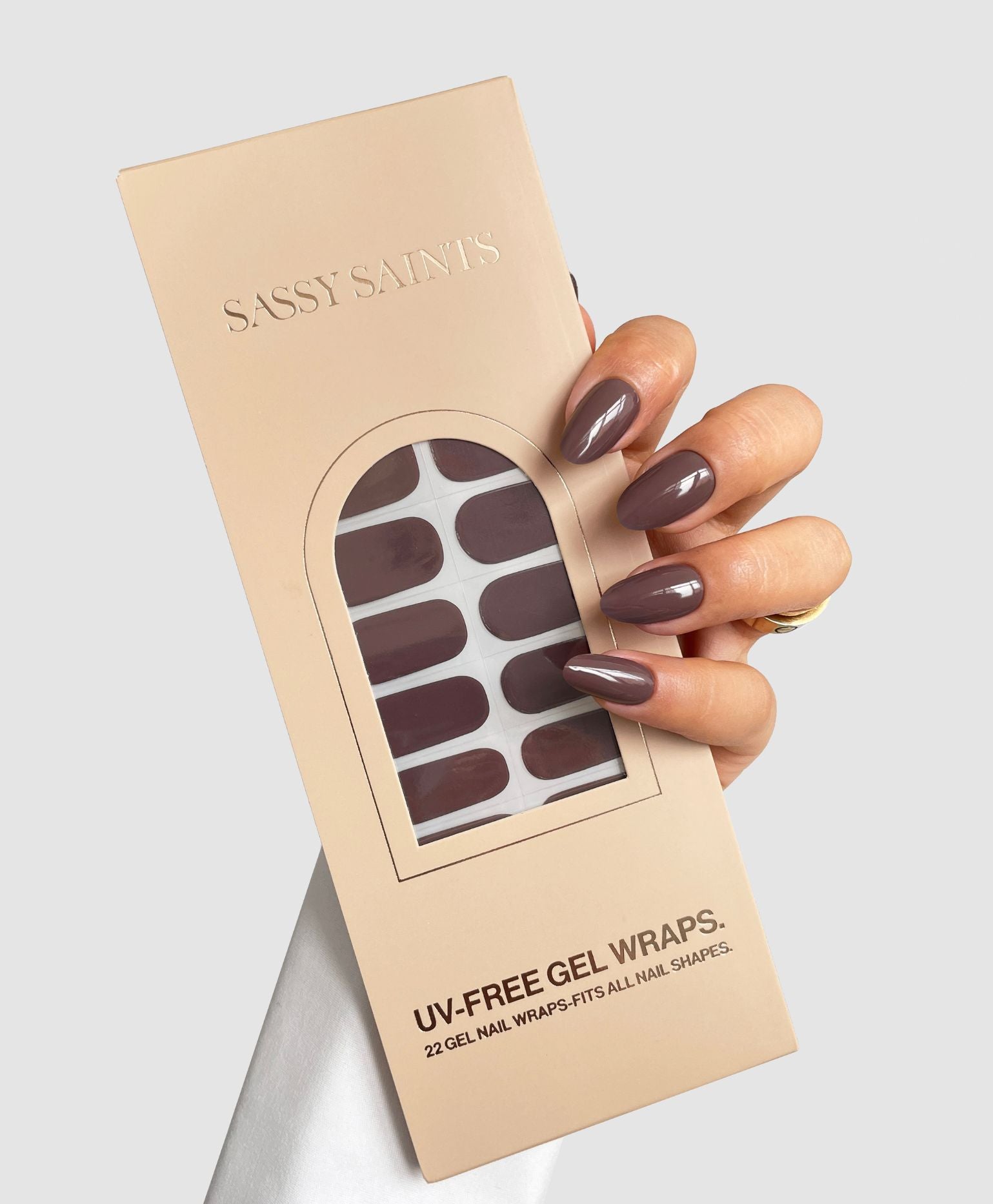 A hand with glossy mauve nails holds a beige box labeled "Sassy Saints – Brings The Salon Home Mocha Blend Gel Wrap," showing matching nail wraps through a front window.