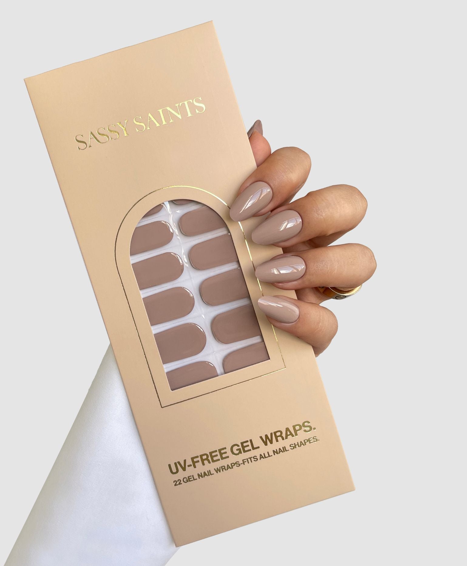A hand with smooth, almond-shaped nude nails holds a beige Sassy Saints - Brings The Salon Home Dusty Stone Gel Wrap box containing 22 nude gel nail wraps. Gold lettering and a window on the packaging reveal the wraps inside.