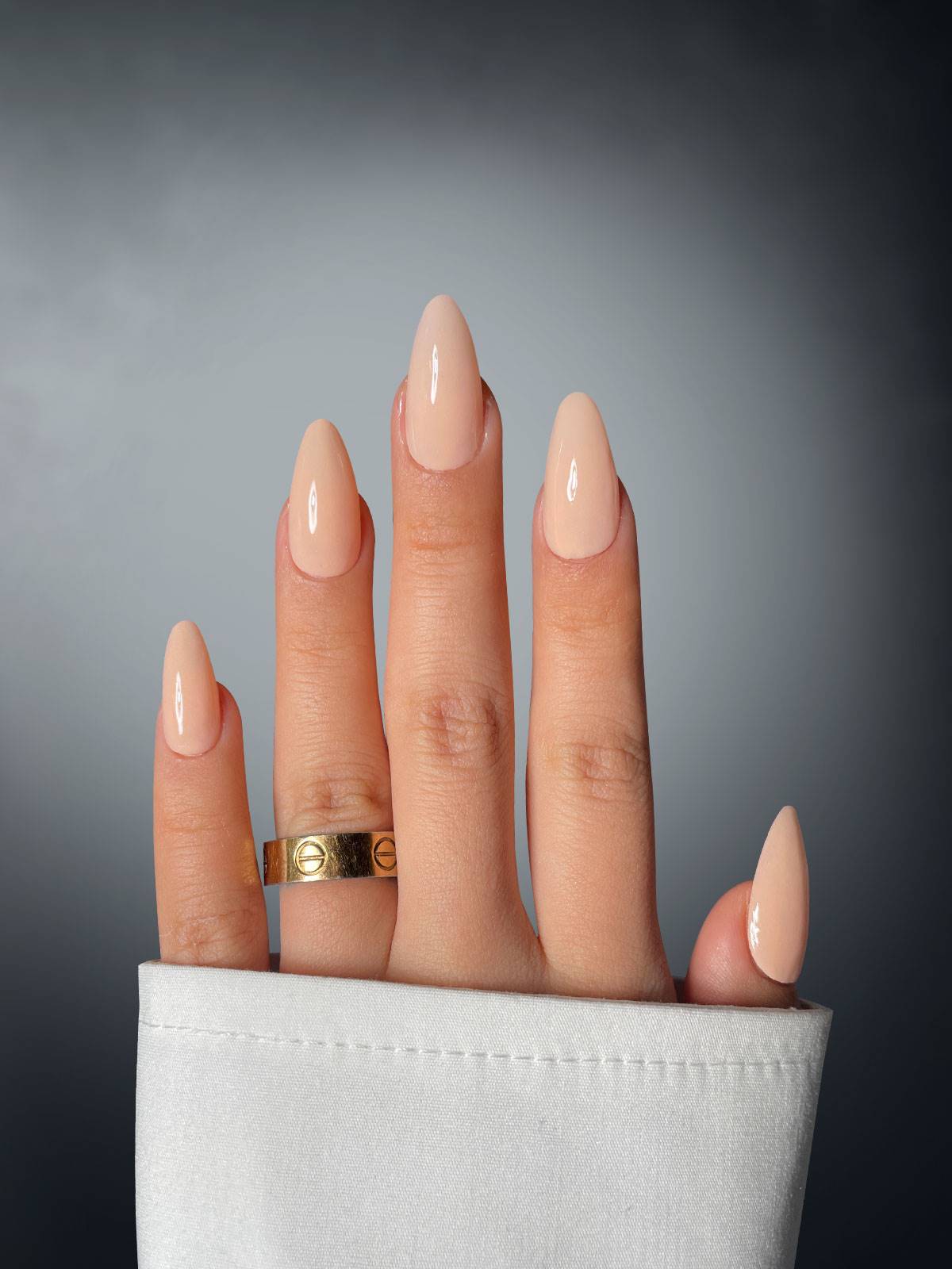 A hand with long, almond-shaped nails coated in Sassy Saints - Brings The Salon Home’s Beige Bombshell Dipping Powder is shown against a gray background. A gold ring adorns the middle finger, while a white sleeve partially covers the wrist.