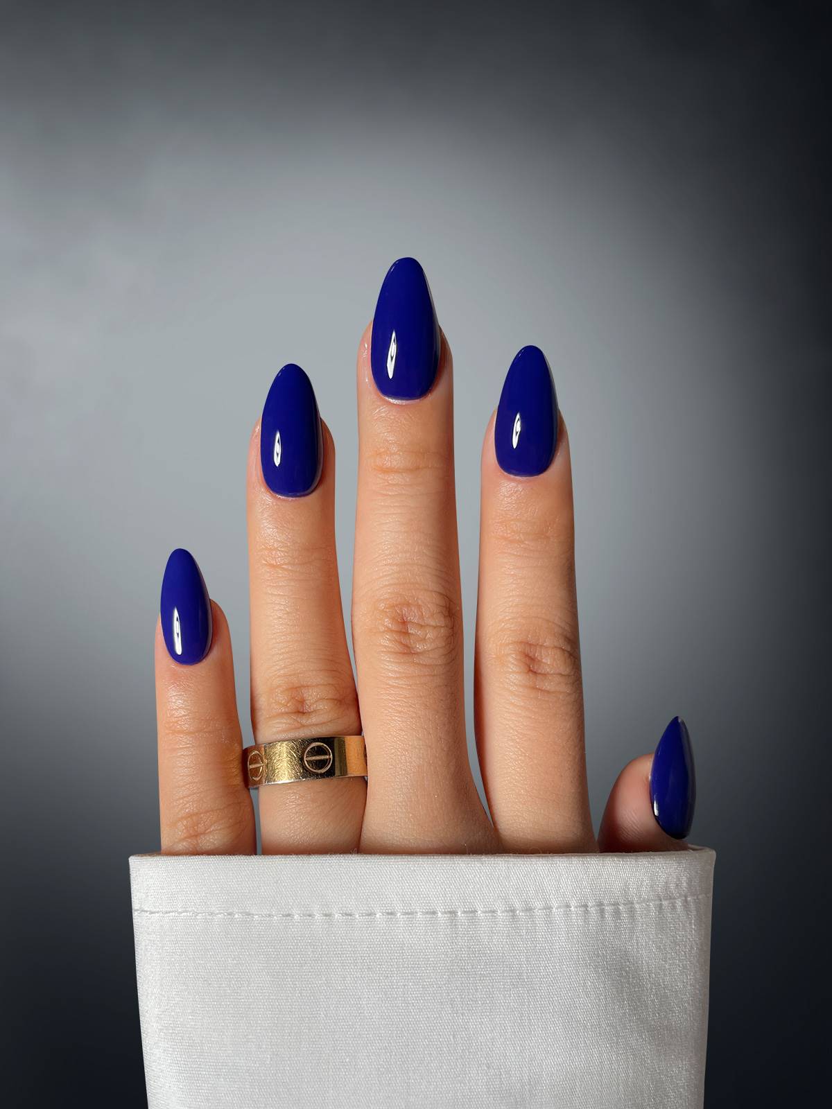 A hand with long, glossy nails in Azure Allure Dipping Powder by Sassy Saints - Brings The Salon Home, adorned with a gold ring, emerges from a white sleeve against a gray gradient background.