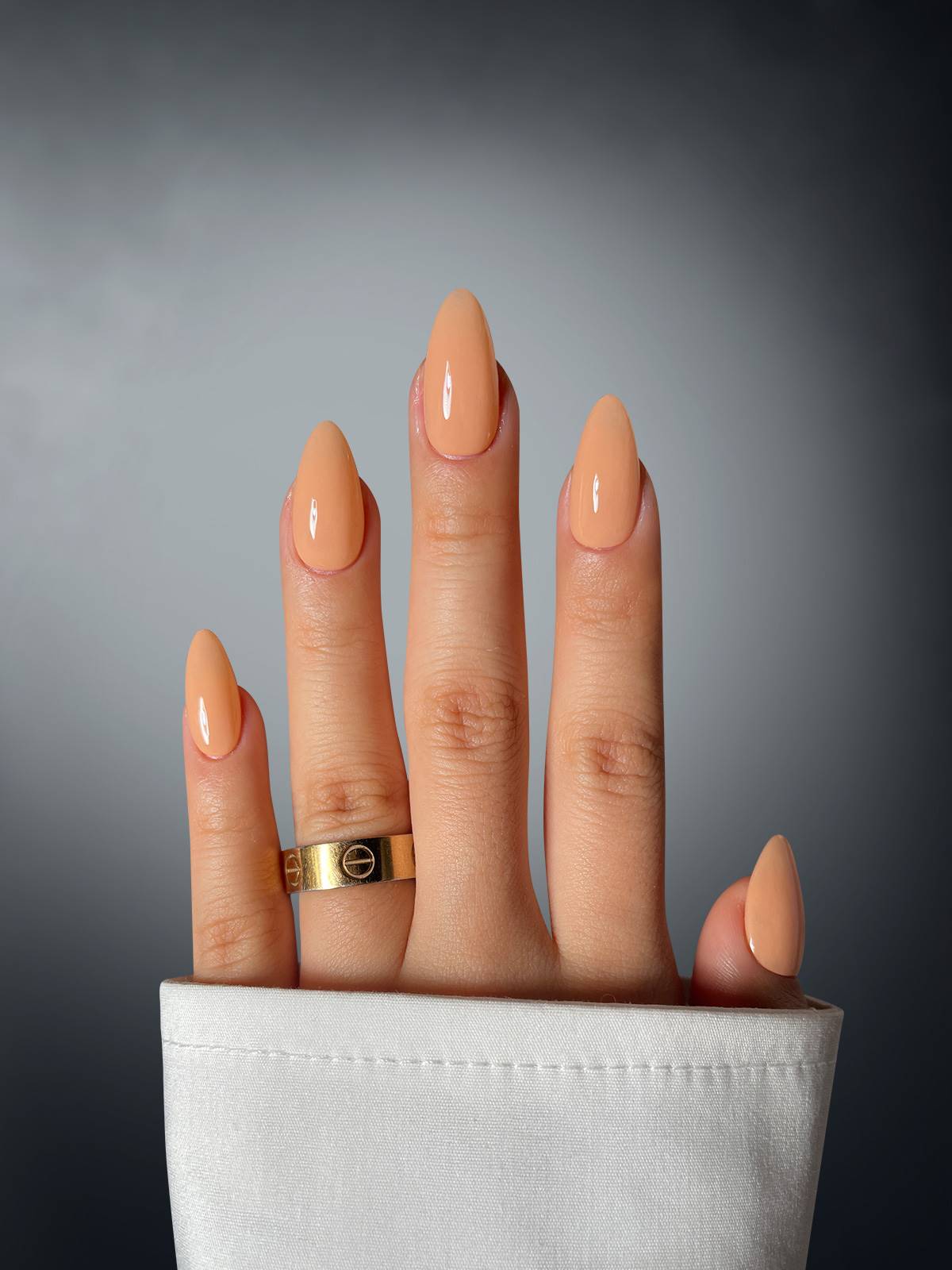 A hand with almond-shaped nude nails in Orange Owned Dipping Powder by Sassy Saints - Brings The Salon Home, wears a gold ring on the middle finger and a white cuffed sleeve, set against a gradient gray background.