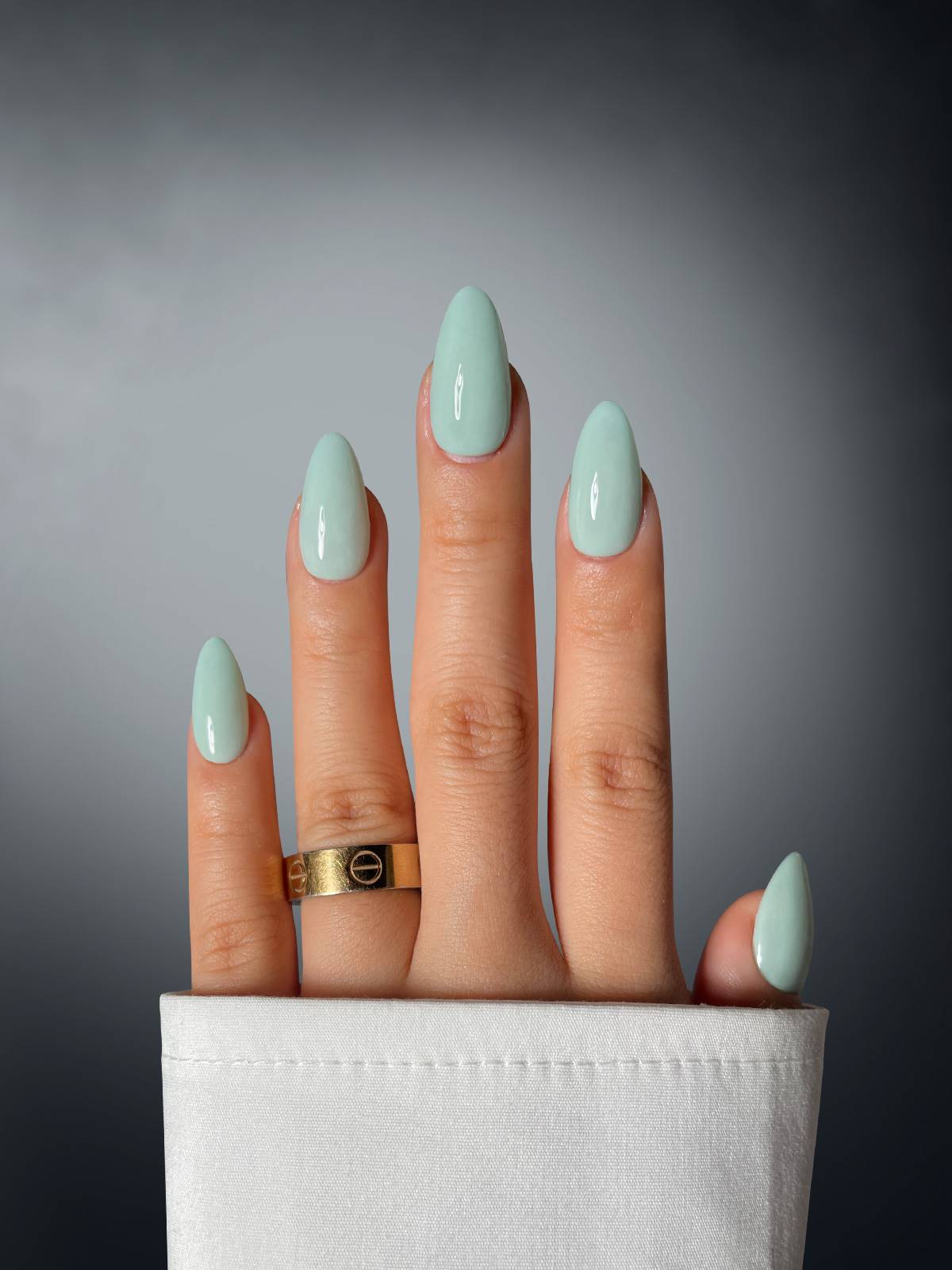 A hand with almond-shaped nails coated in Sassy Saints - Brings The Salon Home's Sage Seduction Dipping Powder poses upright, sporting a gold ring and white sleeve, set against a gray background.