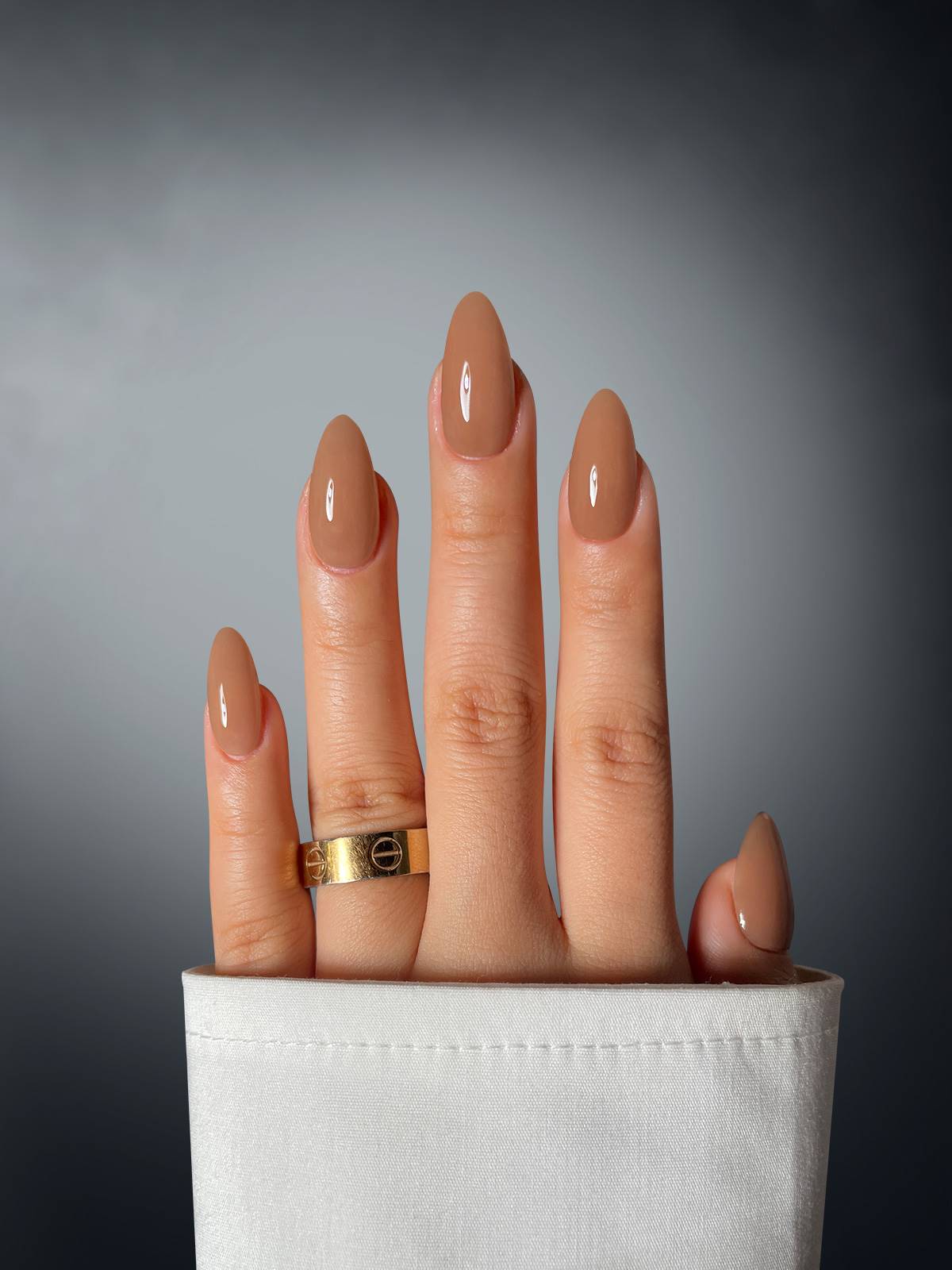 A hand with glossy nude nails polished in Fiery Femme Fatale Dipping Powder by Sassy Saints - Brings The Salon Home, partly covered by a white sleeve, wears a gold ring on the middle finger against a smooth gray gradient background.