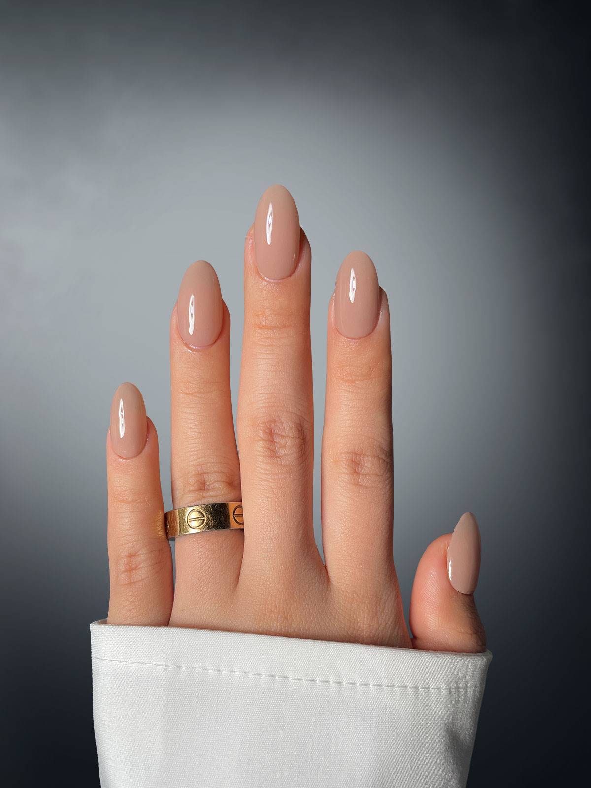 A hand with nude manicured nails in Walnut Wonder Dipping Powder by Sassy Saints, a gold ring on the middle finger, and a white sleeve is shown against a gradient gray background.