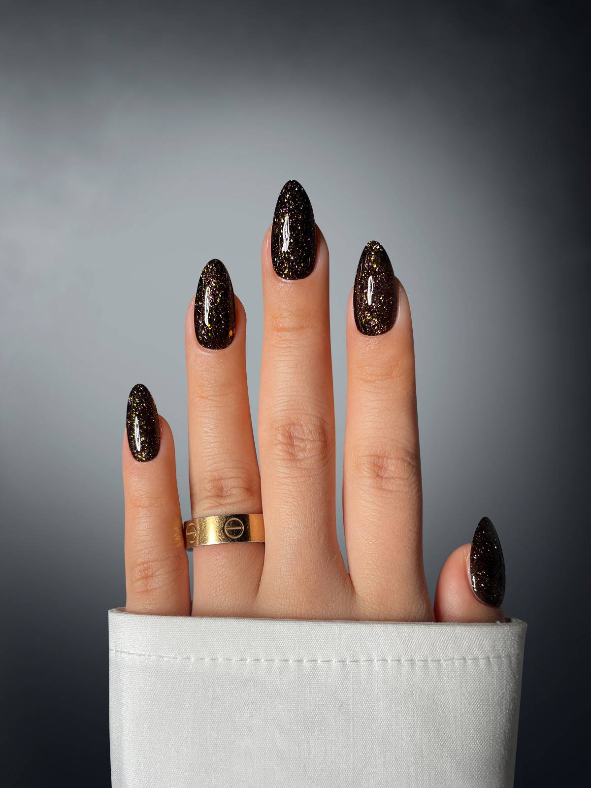 A hand with long, almond-shaped nails showcases Sassy Saints’ Starry Sky Dipping Powder—a black shade with gold glitter. The person wears a white cuffed sleeve and a gold ring on the middle finger against a gradient dark background.