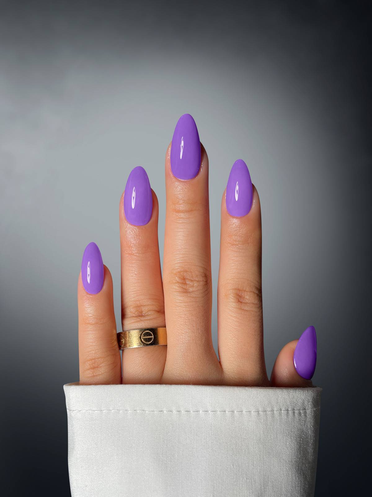 A hand with four fingers, each nail coated in Sassy Saints' Vivacious Violet Dipping Powder, emerges from a white sleeve. The middle finger sports a gold ring—set against a soft gray gradient background.