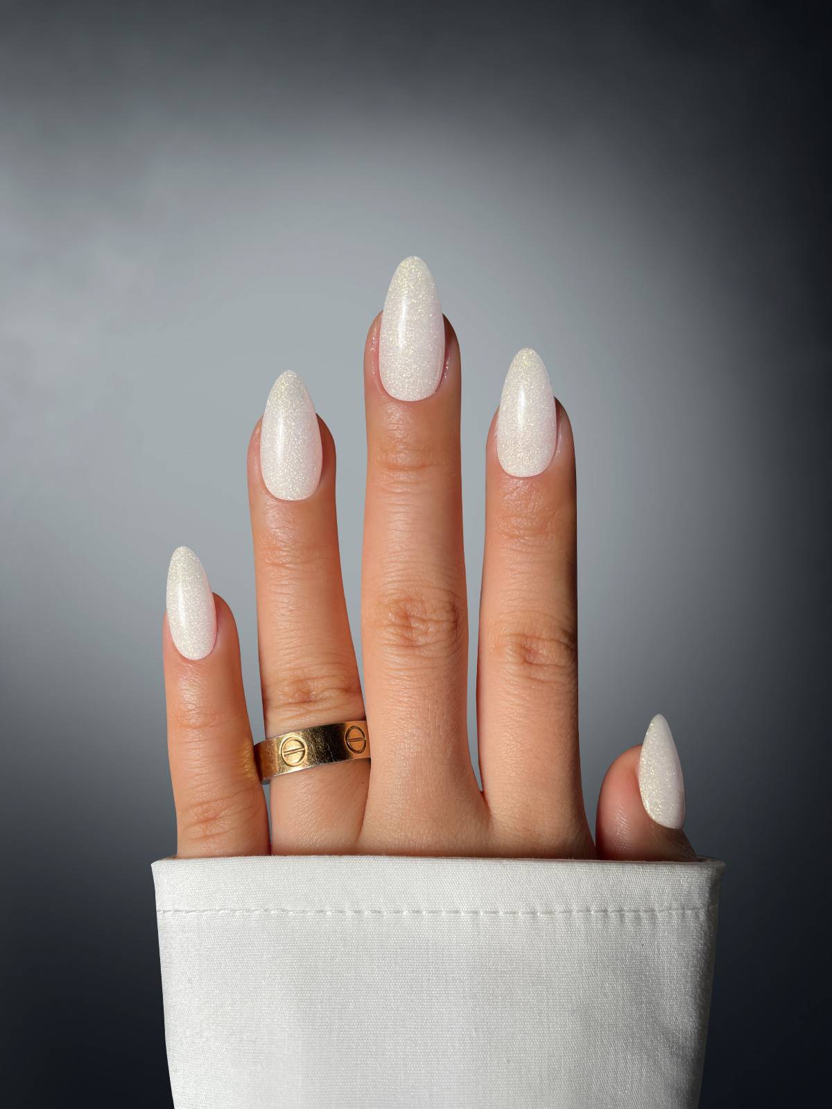 A hand with long, white, almond-shaped nails—created using Sassy Saints Sparkling Queen Dipping Powder—is shown against a dark gradient background. The person wears a gold ring on the middle finger and a white sleeve.