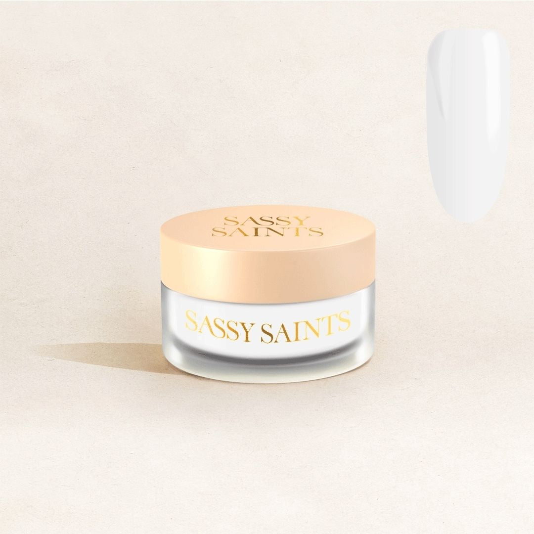 A small frosted glass jar with a beige lid, labeled Sassy Saints – Brings The Salon Home, rests on a light beige surface. The pure white Icy Illusionist Dipping Powder is shown as a glossy nail color swatch in the upper right corner.