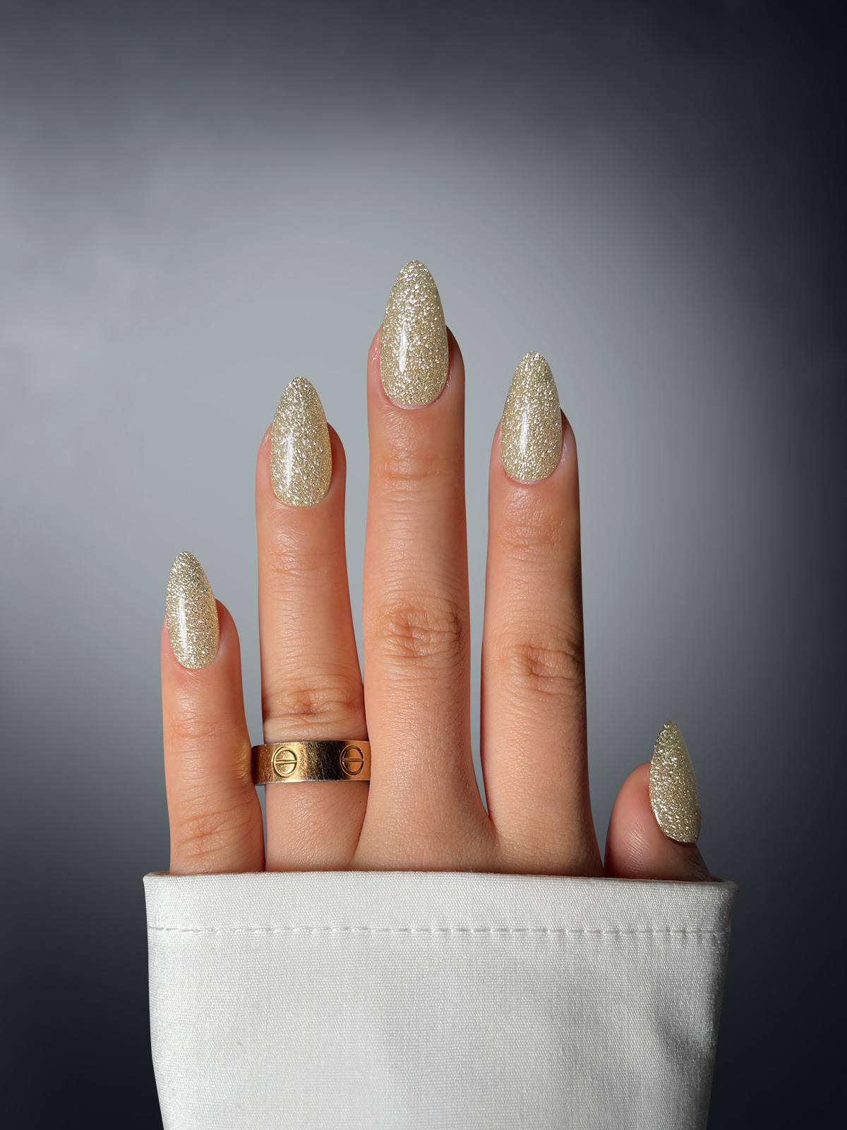 A hand with long, pointed nails painted in Sassy Saints' Champagne Charmer Dipping Powder and adorned with two gold rings on the middle finger is shown against a gray gradient background. The person wears a white long-sleeved shirt.