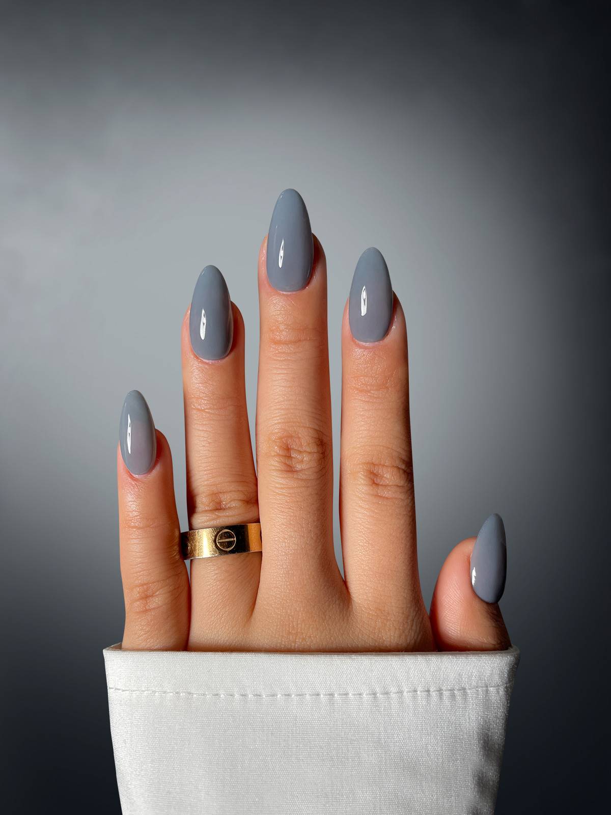 A hand with shiny, gray, almond-shaped nails—created using Sassy Saints’ Crimson Conqueror Dipping Powder—rests against a dark gradient background, accented by a gold ring and a white sleeve at the wrist.