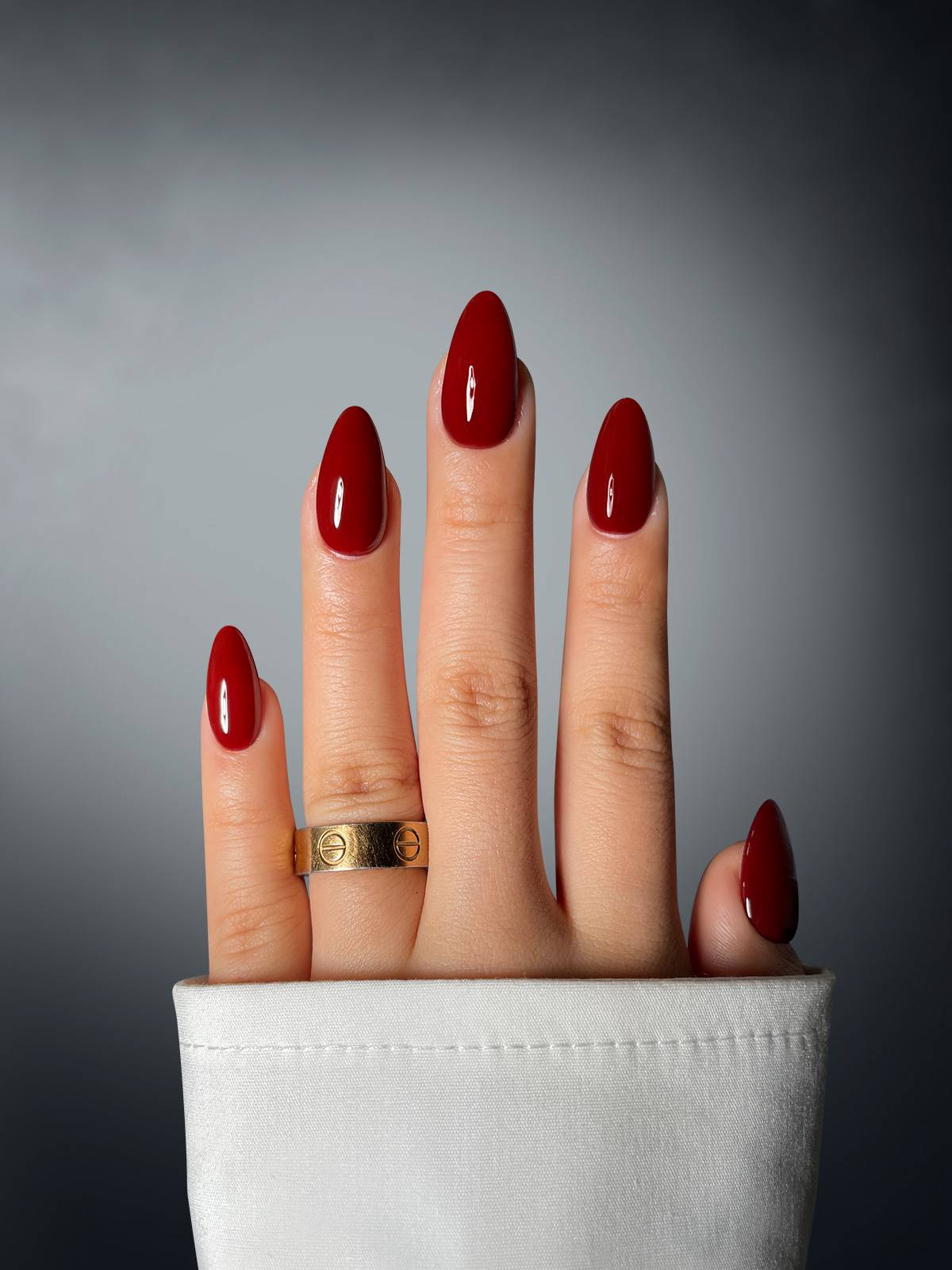 A hand with long, almond-shaped nails in Sassy Saints' Bordeaux Babe Dipping Powder is shown against a gray gradient background, wearing a gold ring on the ring finger and partially covered by a white sleeve.