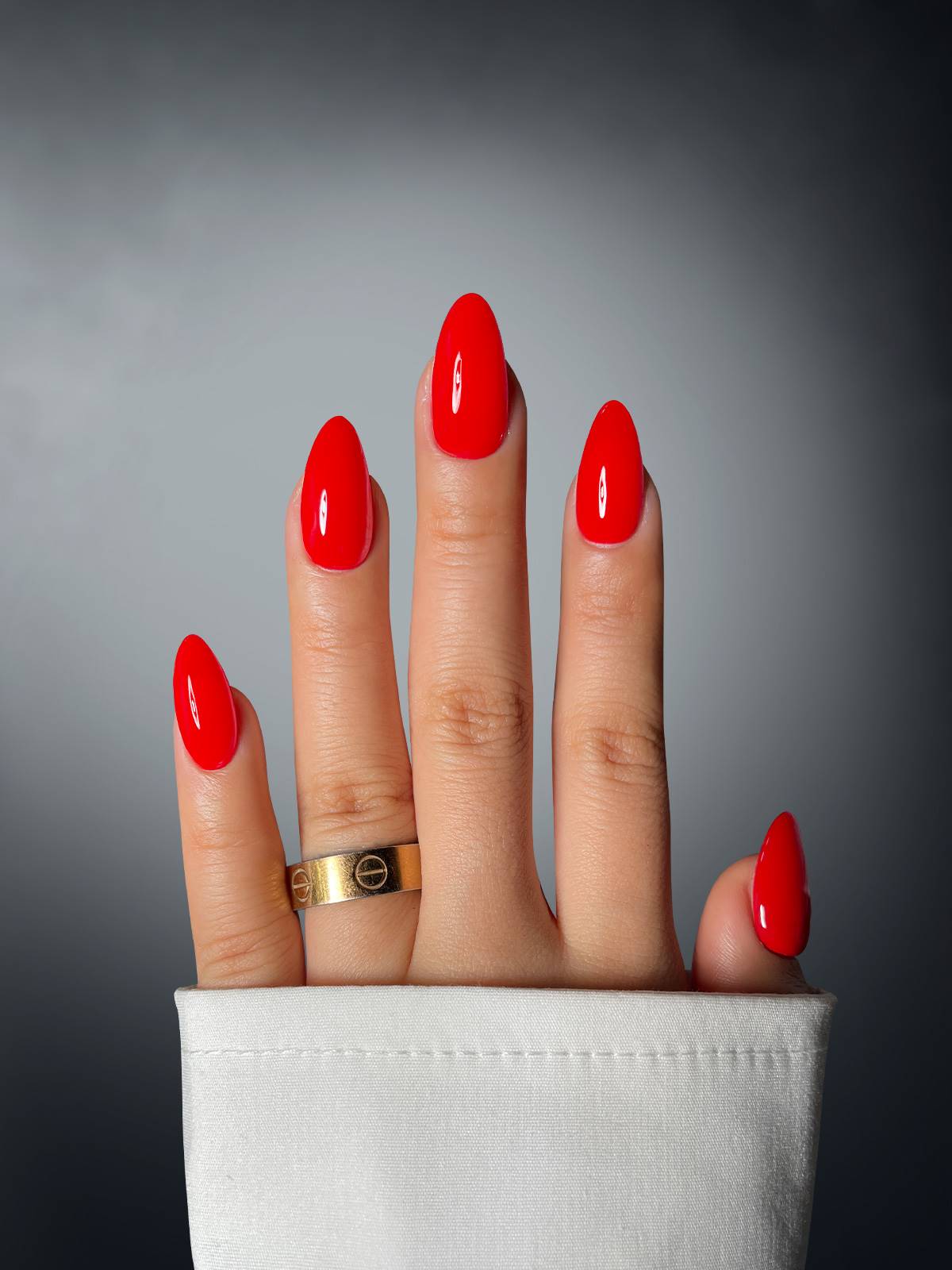 A hand with almond-shaped nails, featuring Sassy Saints' Rouge Romance Dipping Powder, wears a gold ring on the middle finger and is partially covered by a white sleeve against a gray gradient background.