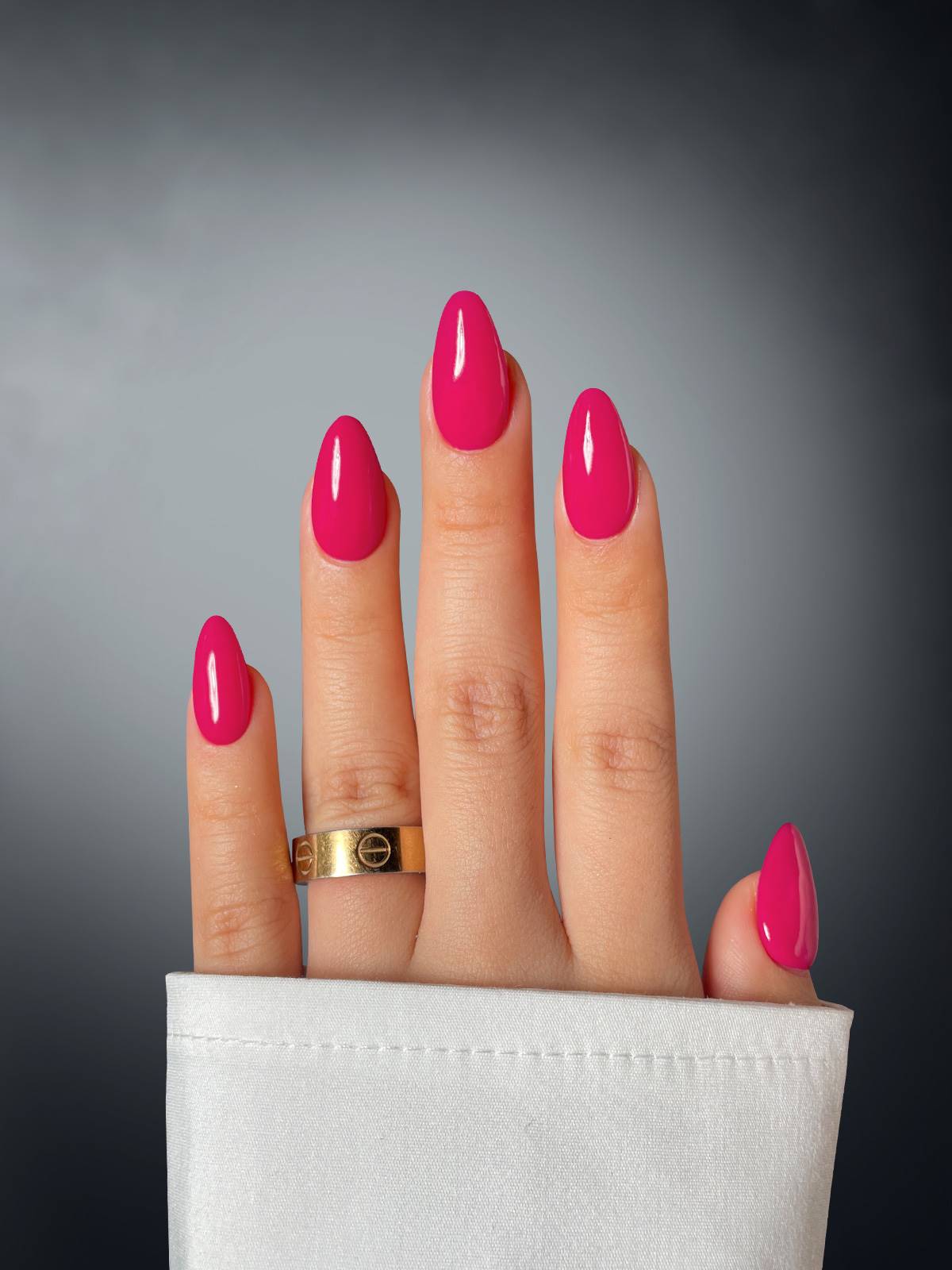A hand with almond-shaped nails in Ravishing Rose by Sassy Saints - Brings The Salon Home, wearing a gold ring and partially covered by a white sleeve, is shown against a dark gradient background.