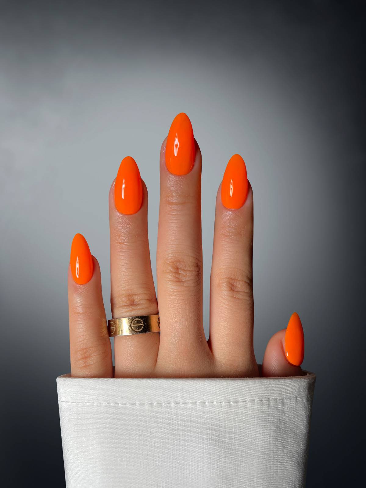 A hand with long, glossy, bright orange almond-shaped nails in Pumpkin Posh Dipping Powder by Sassy Saints - Brings The Salon Home is shown against a gray gradient background. The person wears a gold ring and a white cuffed sleeve.