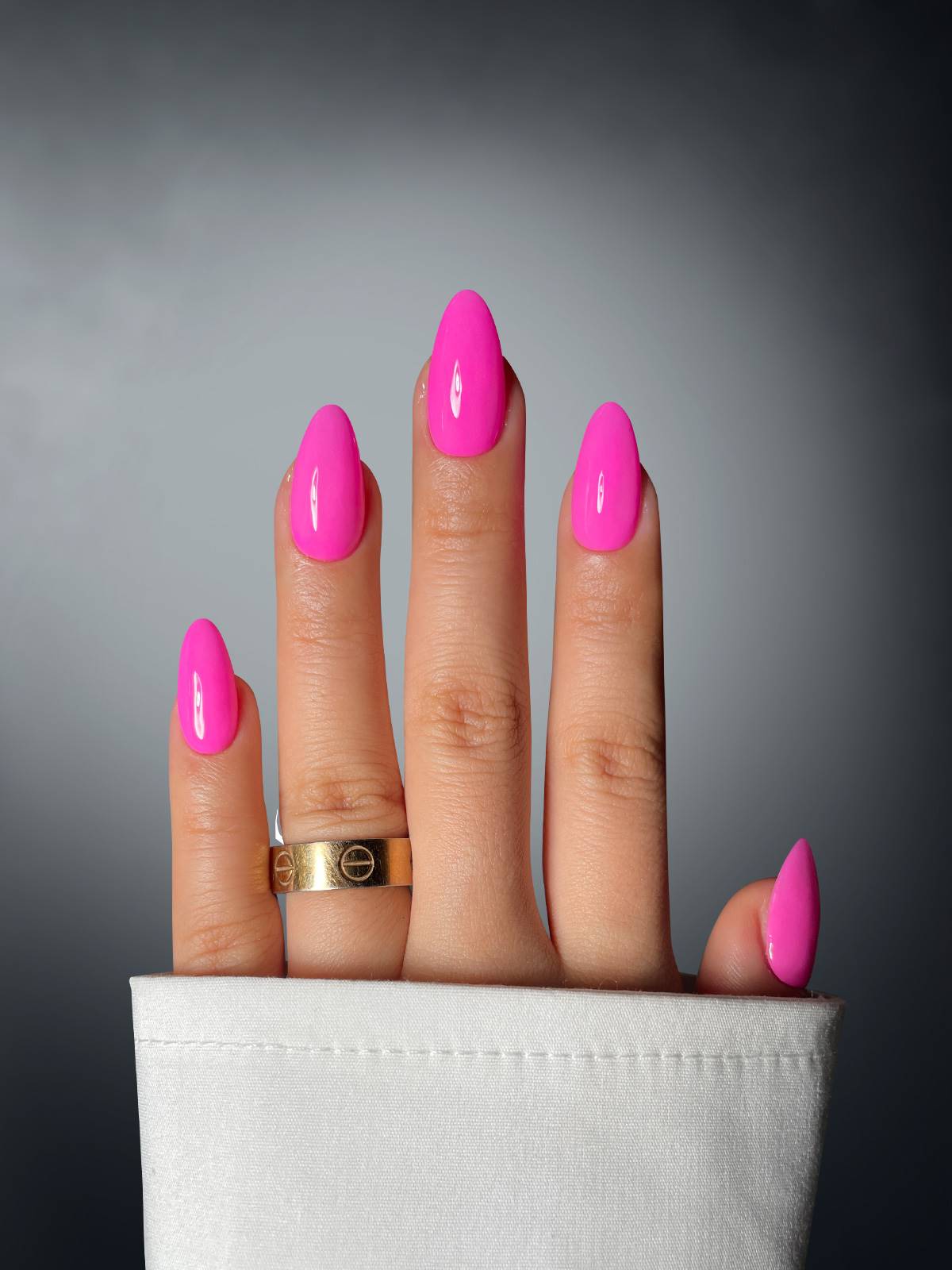 A hand with four fingers, almond-shaped nails in Sassy Saints - Brings The Salon Home’s Orchid Obsession Dipping Powder, and a gold ring emerges from a white sleeve against gray; the middle two fingers are unusually close together.