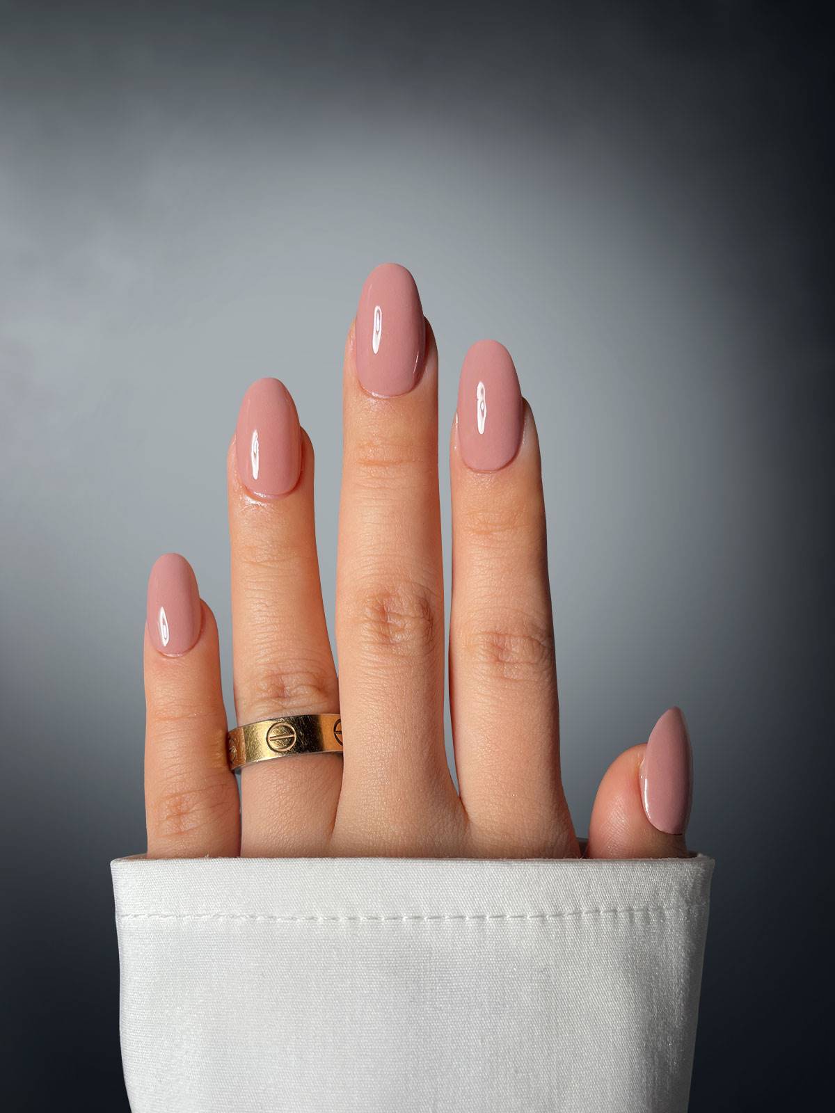 A hand with four pink-polished fingers, featuring Taupe Temptress Dipping Powder by Sassy Saints - Brings The Salon Home, displays a gold ring on the middle finger. The hand extends from a white sleeve against a soft grey gradient background.