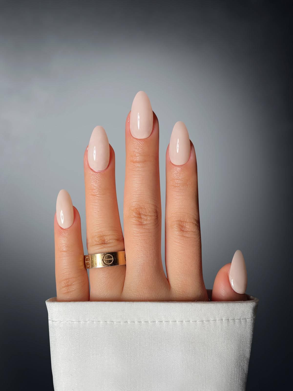 A hand with almond-shaped nude nails, styled with Sassy Saints' French Flair Dipping Powder, wears a gold ring on the middle finger. The white shirt sleeve and gray gradient background complete the elegant look.