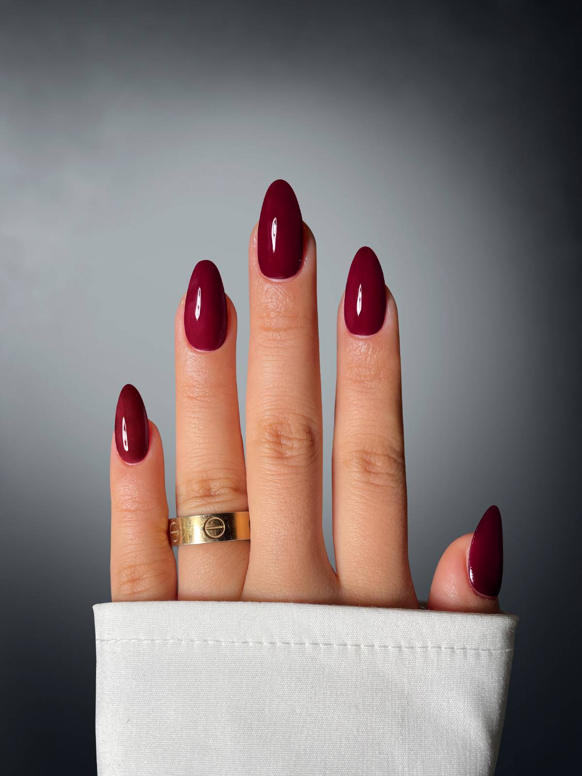 A hand with burgundy oval nails, created using Mink Maven Dipping Powder by Sassy Saints - Brings The Salon Home, and a gold ring on the middle finger is shown against a gradient background, partially covered by a white sleeve.