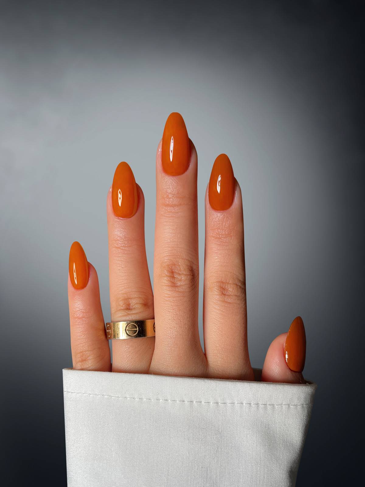 A hand with long, glossy, orange almond-shaped nails—created using Toffee Temptation Dipping Powder by Sassy Saints—is shown against a gradient gray background, accessorized with a gold ring and white cuffed sleeve.