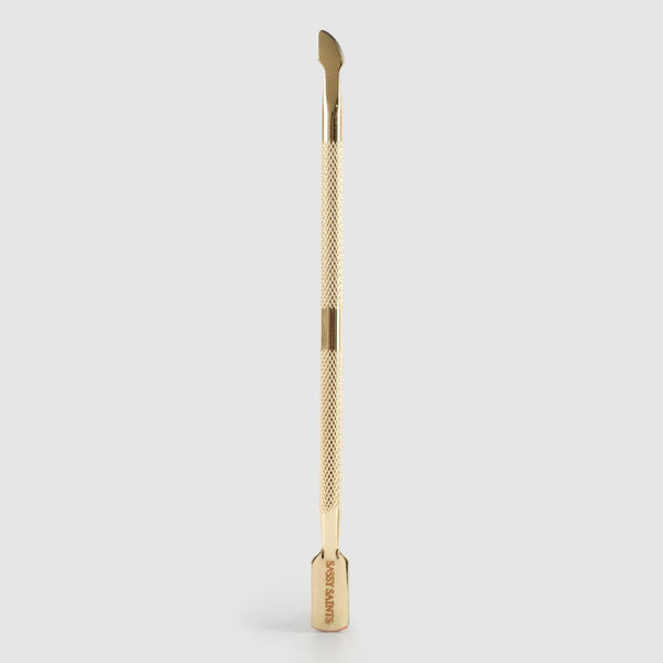 The Sassy Saints Gold Cuticle Pusher features a textured grip and dual-ended tips for manicure prep, shown standing upright against a plain light gray background. Brings The Salon Home.