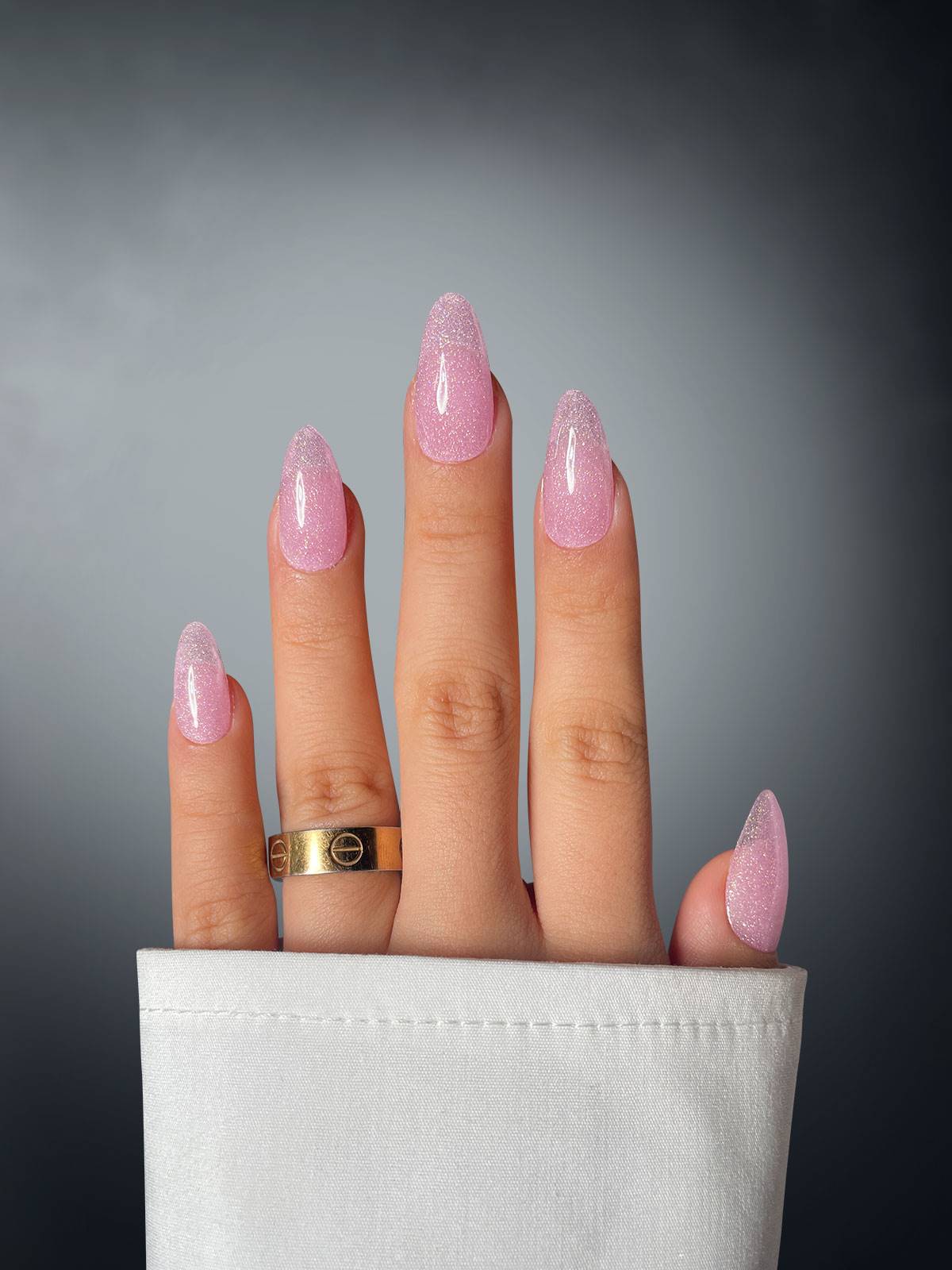 A hand with four fingers shows off long, almond-shaped nails in glittery pink—Shimmering Siren Dipping Powder by Sassy Saints. A gold ring shines on the middle finger, and a white shirt sleeve covers the wrist against a smooth gray background.