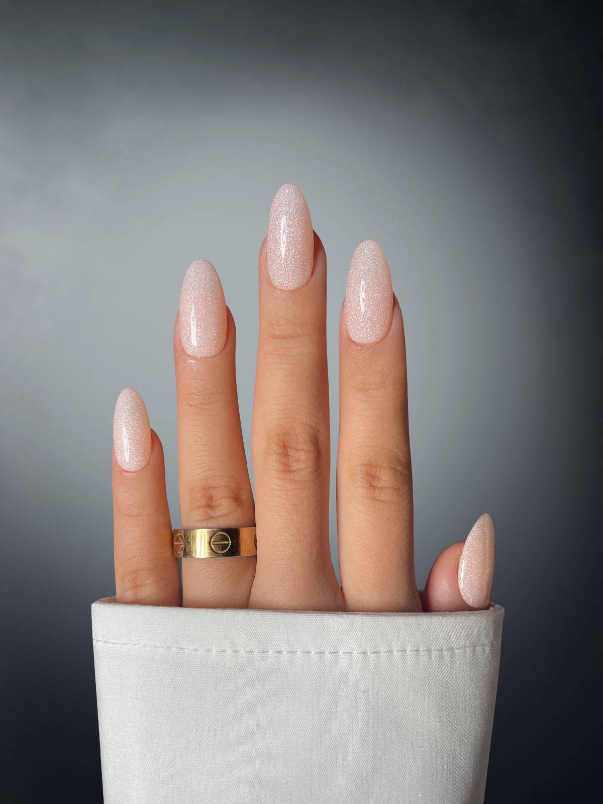 A hand with long, almond-shaped nails in pale pink glitter using Sassy Saints Sparkle Suede Dipping Powder, plus a gold ring on the middle finger, is shown against a gray background, partially emerging from a white sleeve.