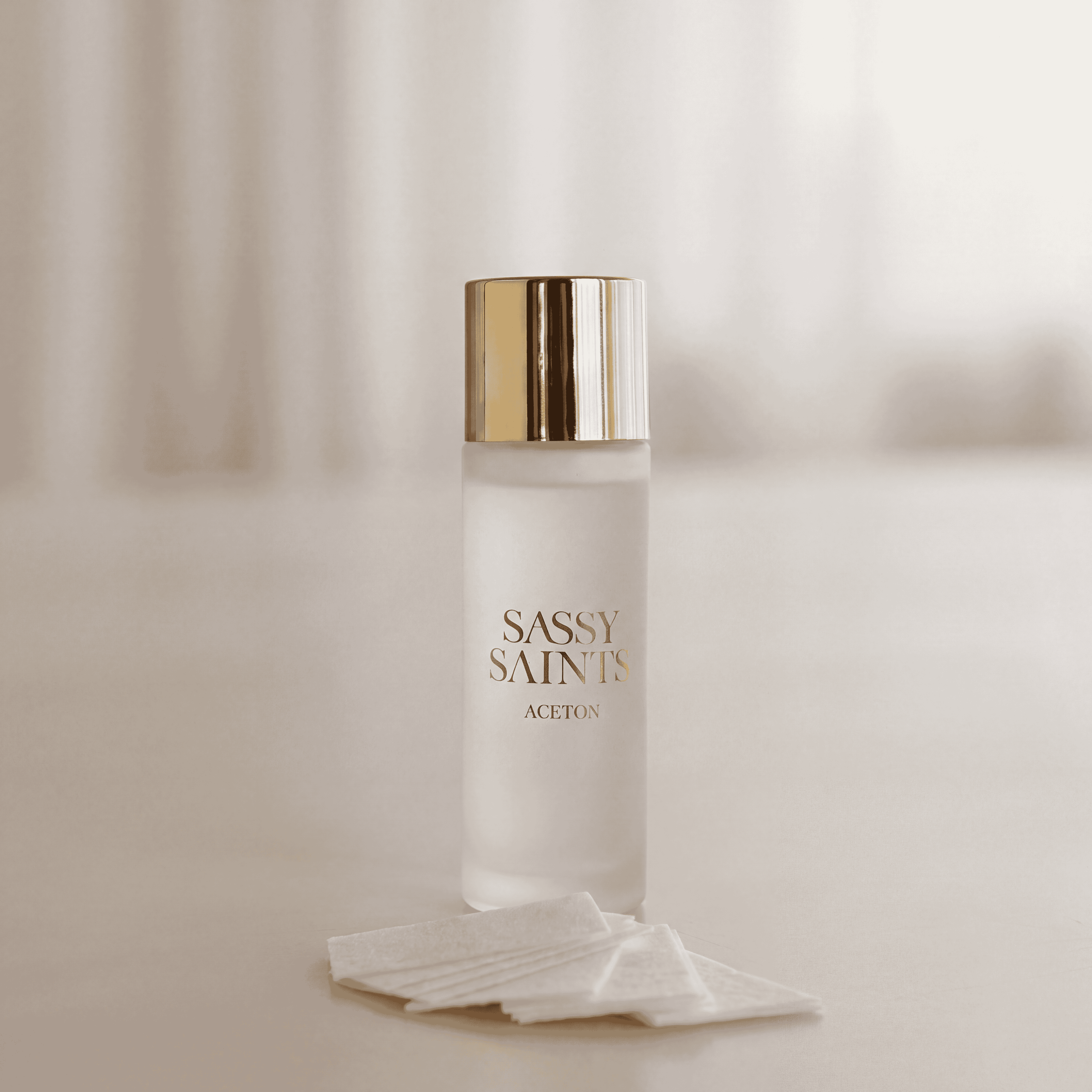 A frosted glass bottle of Sassy Saints Remover set - 450ml with a gold cap, accompanied by a small stack of white cotton pads.