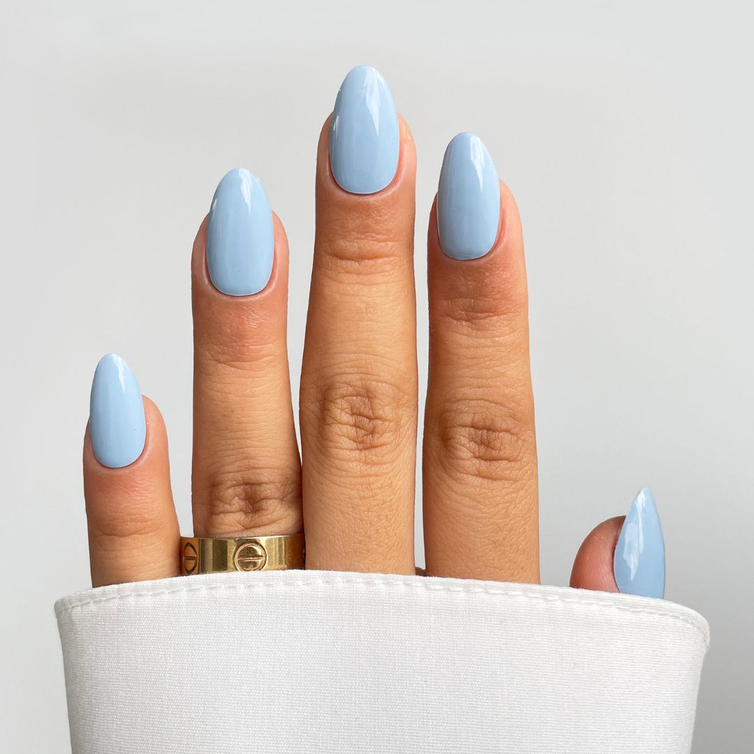 A hand with almond-shaped nails in Sassy Saints' Aqua Clouds Gel Wrap, light blue, wears a gold ring on the middle finger and is partly covered by a white sleeve against a plain background.