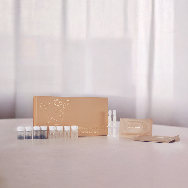 A tan box labeled Bio-Microneedling Facial Kit by Sassy Saints - Brings The Salon Home rests on a white surface, with small bottles, ampoules, and packets neatly arranged beside it against a softly lit backdrop with sheer curtains.