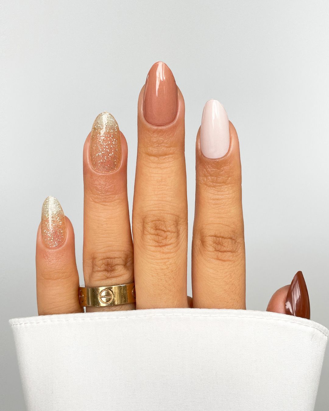 A hand with Sassy Saints' Bare Minimum Gel Wraps—two gold glitter, one nude pink, one pale pink, and one brown nail—is shown with a gold ring and white sleeve against a light gray background. Brings The Salon Home.