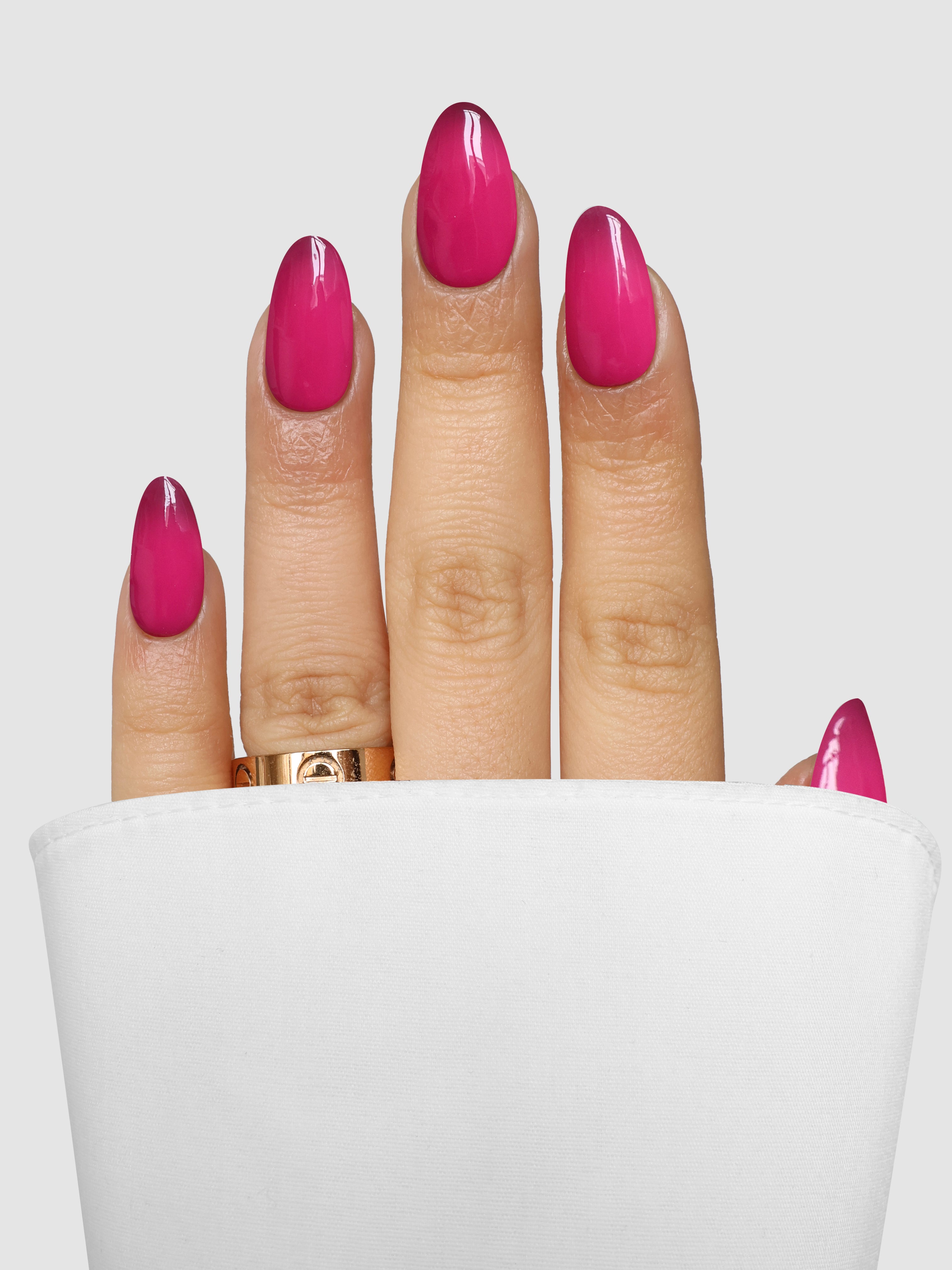 A hand with shiny pink nails, colored with Sassy Saints - Brings The Salon Home's Berry Blush Dipping Powder, and a gold ring is partially covered by a white fabric cuff against a plain light gray background.