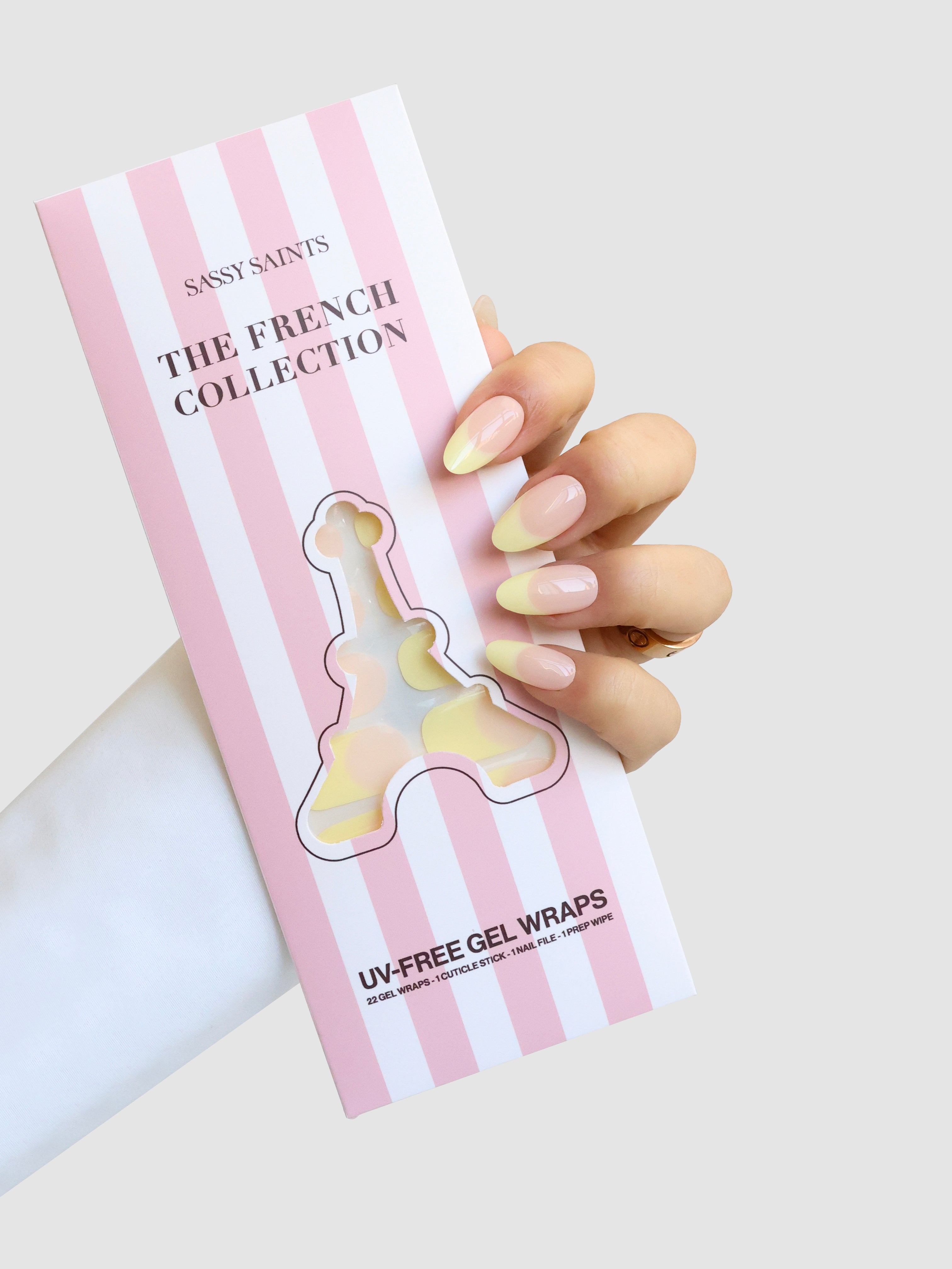 A hand with glossy, light pink and yellow French tip nails holds a pink and white striped box labeled Sassy Saints - Brings The Salon Home's Beurre Doux, featuring an Eiffel Tower-shaped window.