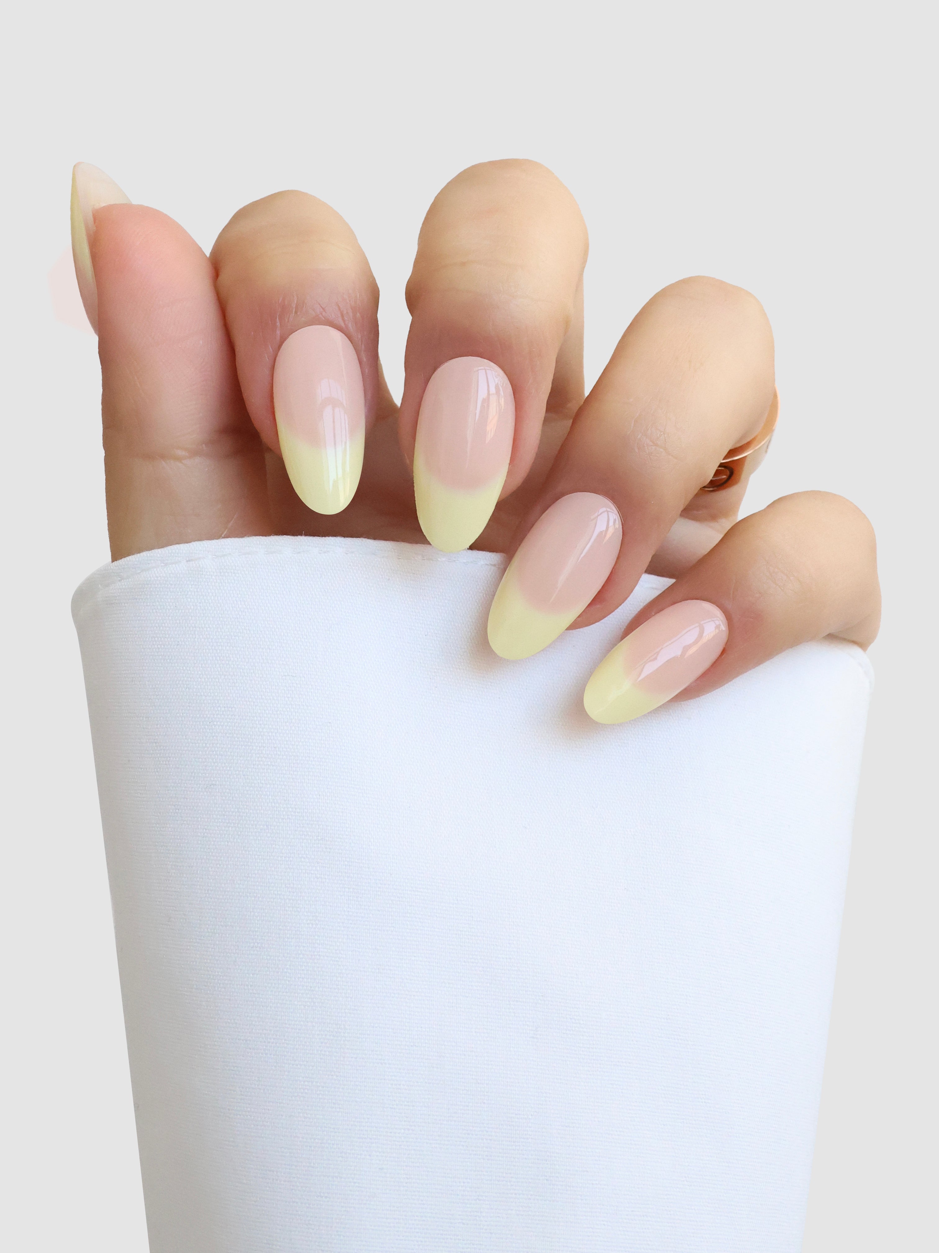 A hand with almond-shaped nails showcases Sassy Saints' Beurre Doux pastel yellow French tips, partially covered by a white sleeve against a light grey background.