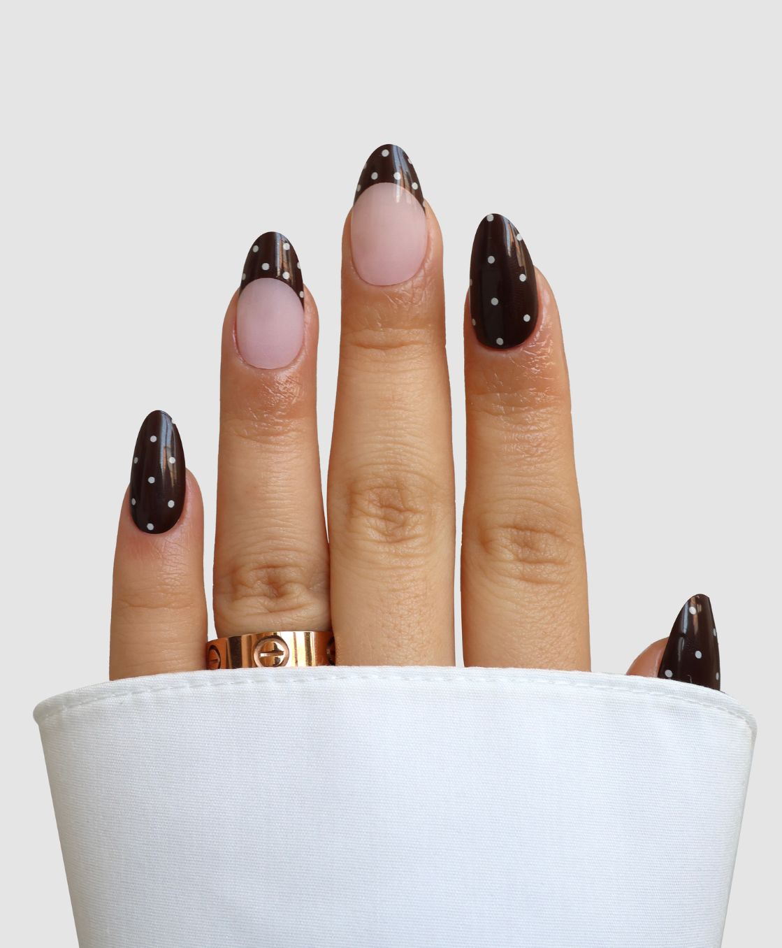 A hand models Sassy Saints' Dark Side Darling Gel Wrap: almond nails with dark brown polish, white polka dots, nude cuticle base, a gold ring on the finger, and a white sleeve partially covering the hand.