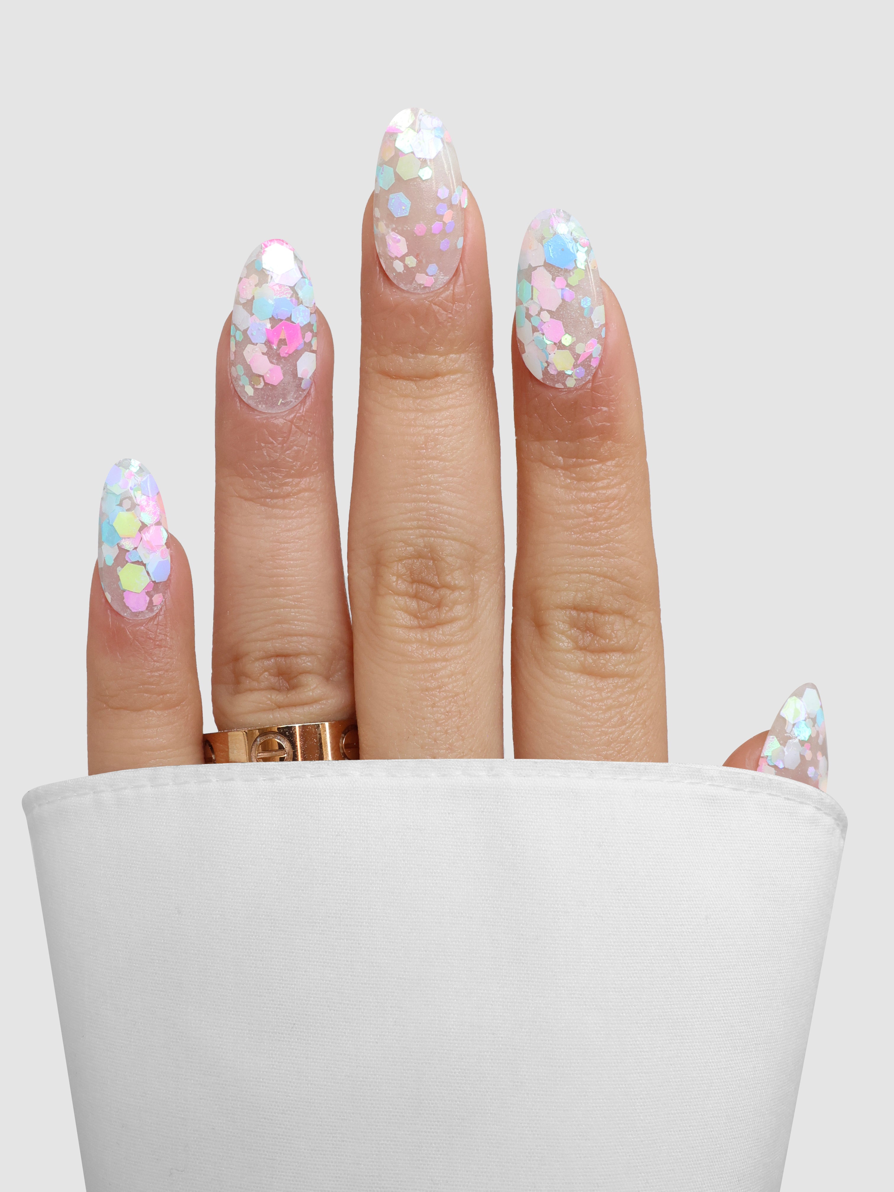 A hand with long, oval nails shows off pastel and iridescent glitter from Sassy Saints' Bloom Sprinkle Dipping Powder, partially covered by white fabric. A gold ring adorns the middle finger, set against a light gray background.