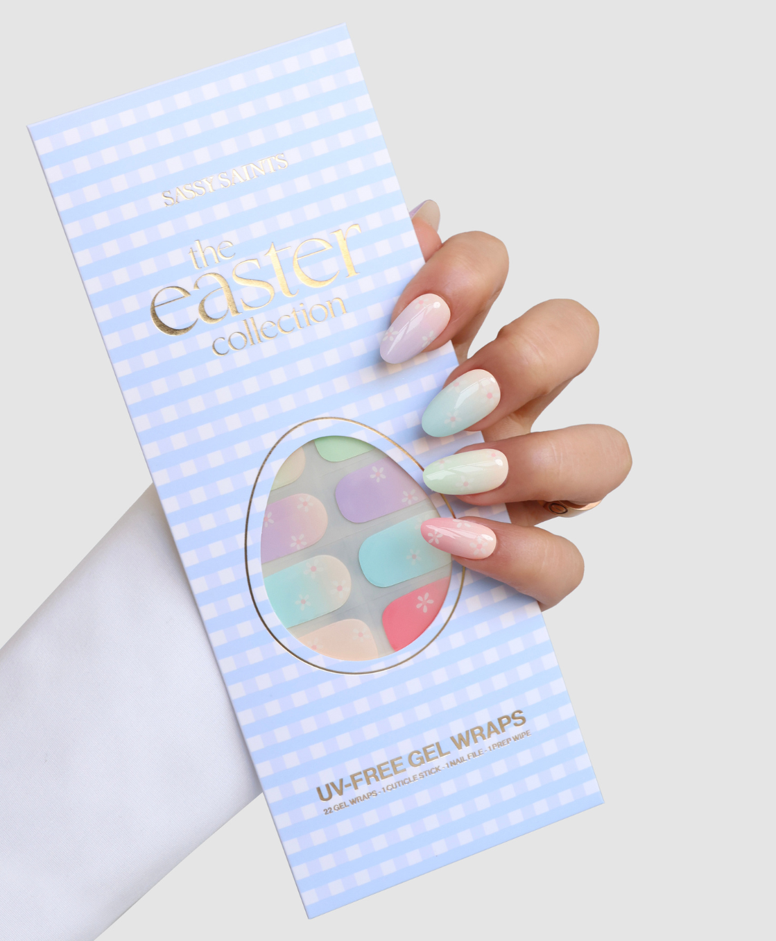 A hand with pastel ombre nails holds a package labeled Blooming Days by Sassy Saints - Brings The Salon Home, featuring pastel nail wraps with small floral designs on a blue gingham background.