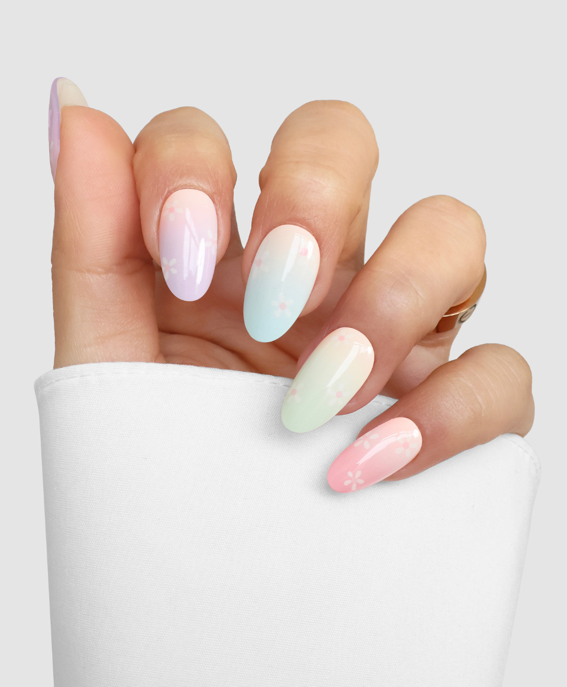 A hand with long almond-shaped nails wears Sassy Saints - Brings The Salon Home's Blooming Days: pastel gradient nails with white flower accents, set against a light background. The person sports a white sleeve.