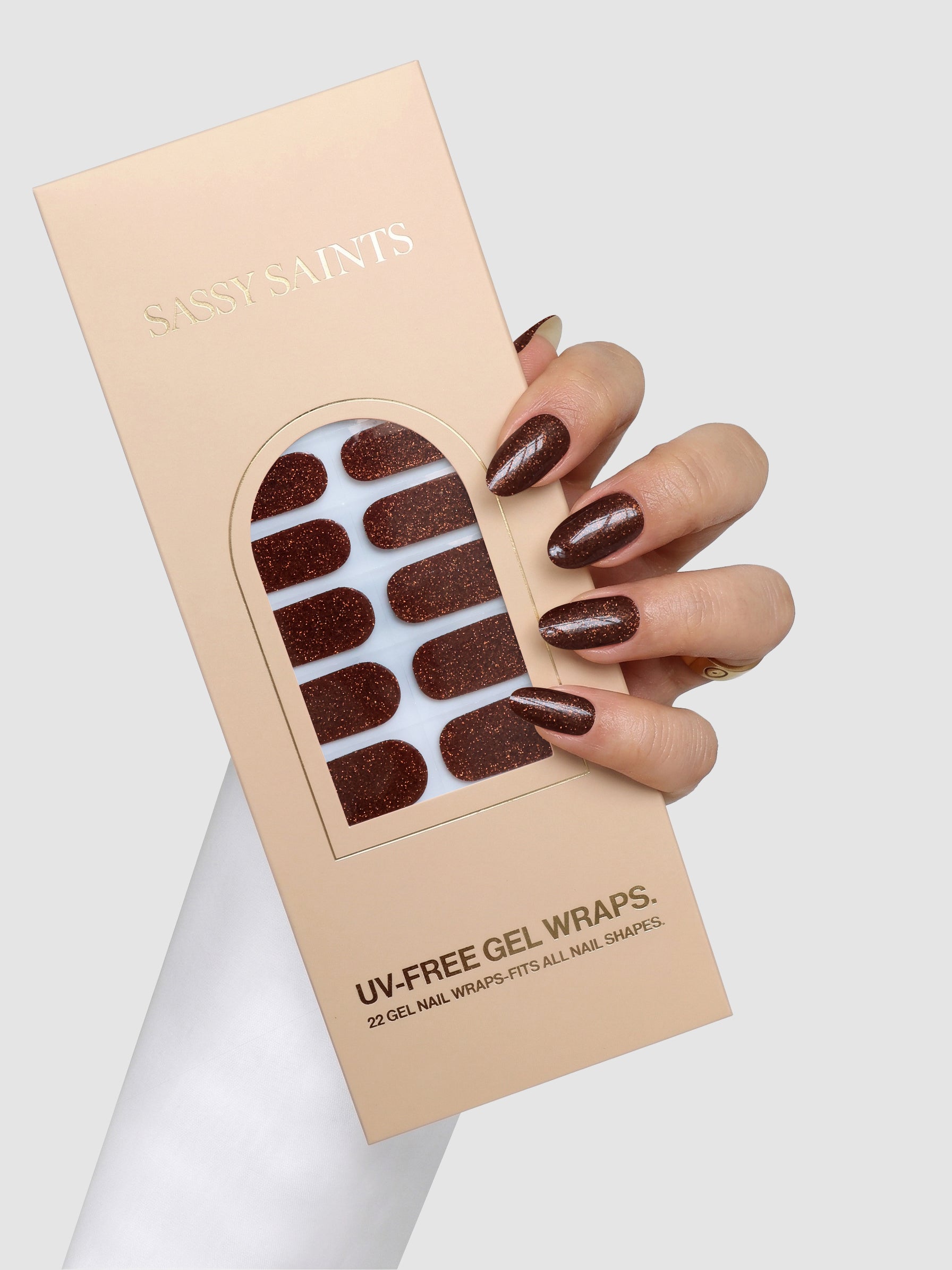 A hand with sparkly brown gel nails holds a box of Sassy Saints - Brings The Salon Home Bronze Behaviour Gel Wraps, featuring matching glittery nail wraps inside the packaging.