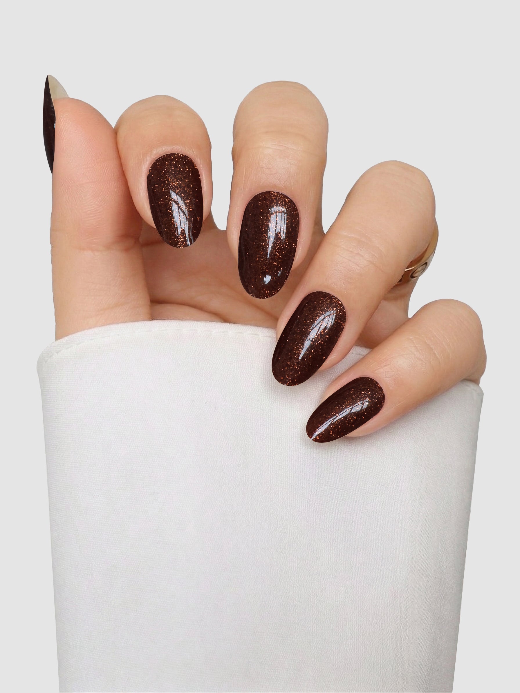 A hand with long, almond-shaped nails features Sassy Saints - Brings The Salon Home’s Bronze Behaviour Gel Wrap—a glossy, dark brown polish with subtle glitter—displayed against a light gray background. The hand is partially covered by a white sleeve.
