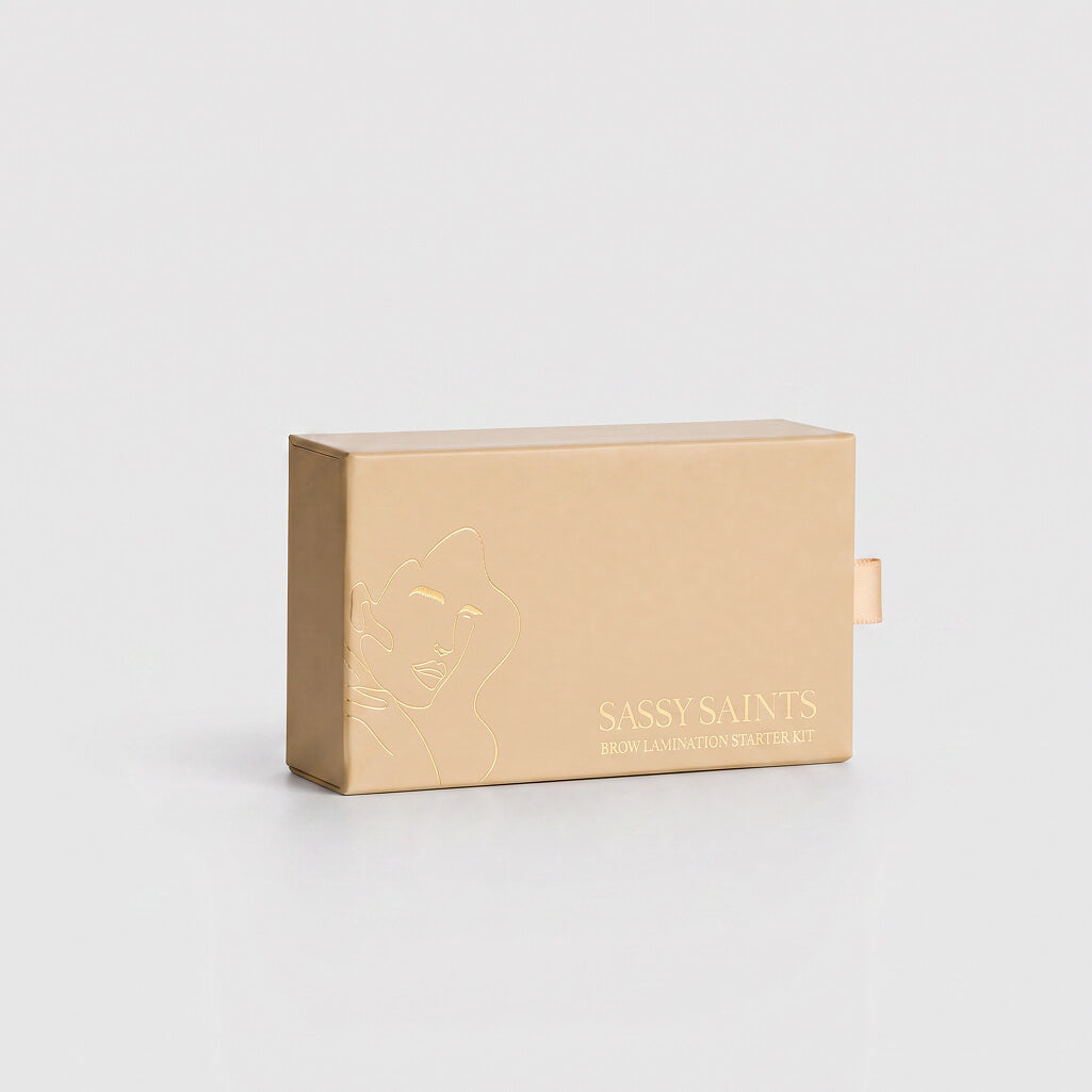 A beige rectangular box labeled “Brow Lamination Mini Kit” by Sassy Saints - Brings The Salon Home, featuring a minimalist line drawing of a face on the left side, designed for easy at-home use and displayed on a plain light background.
