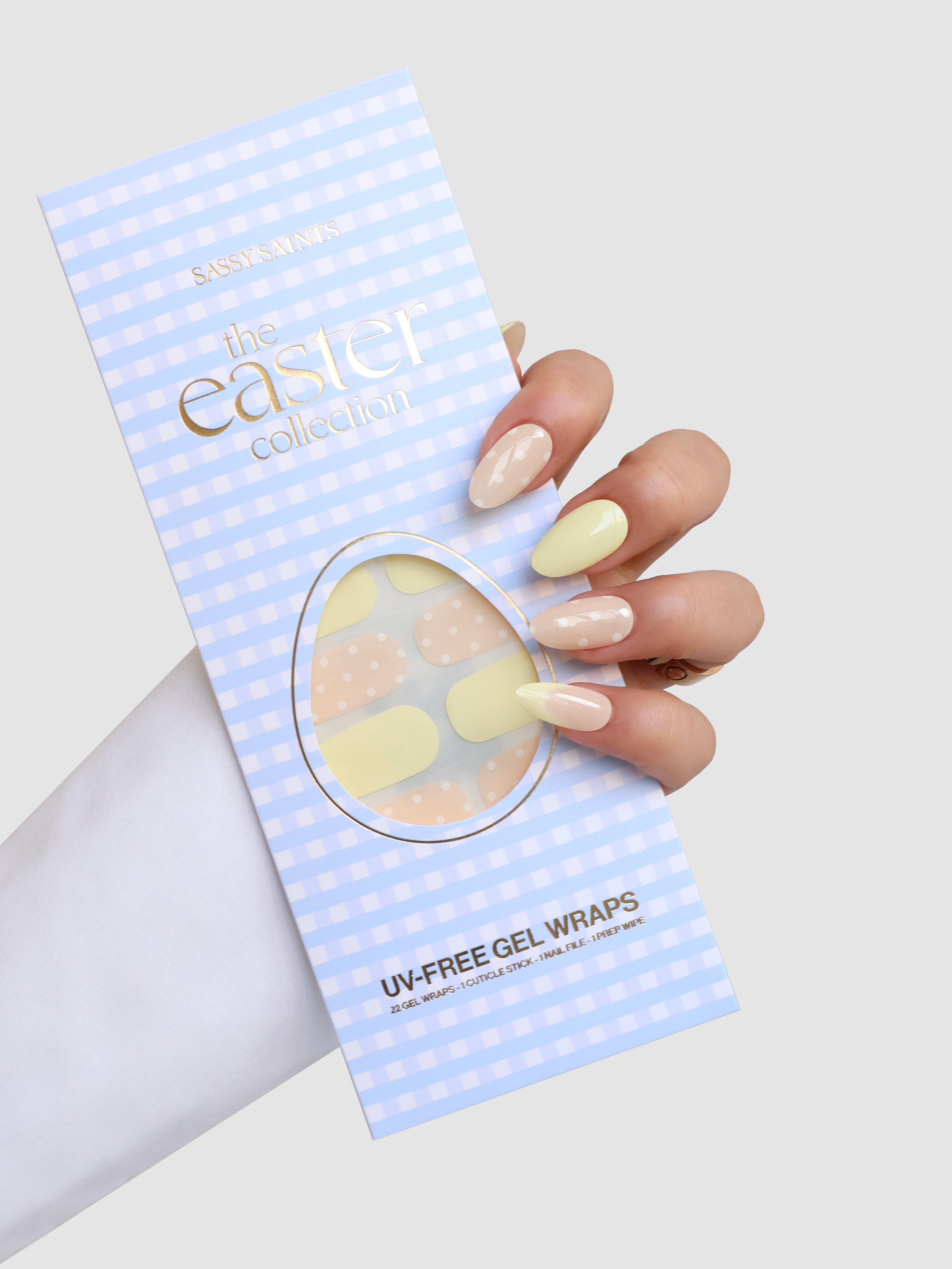 A hand with pastel yellow and beige Easter-themed nails holds a blue checkered package labeled "Buttercup Kiss" by Sassy Saints - Brings The Salon Home, featuring coordinating gel nail wraps for the Easter season.