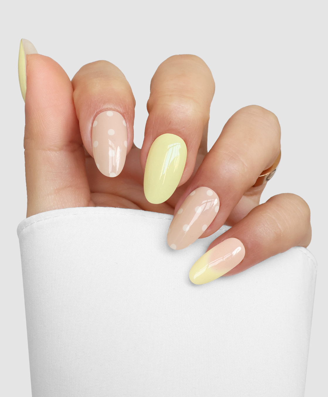 A hand with Sassy Saints' Buttercup Kiss almond nails shows a pastel yellow and nude palette—some nails solid yellow, others nude with white polka dots—partially covered by a white sleeve.