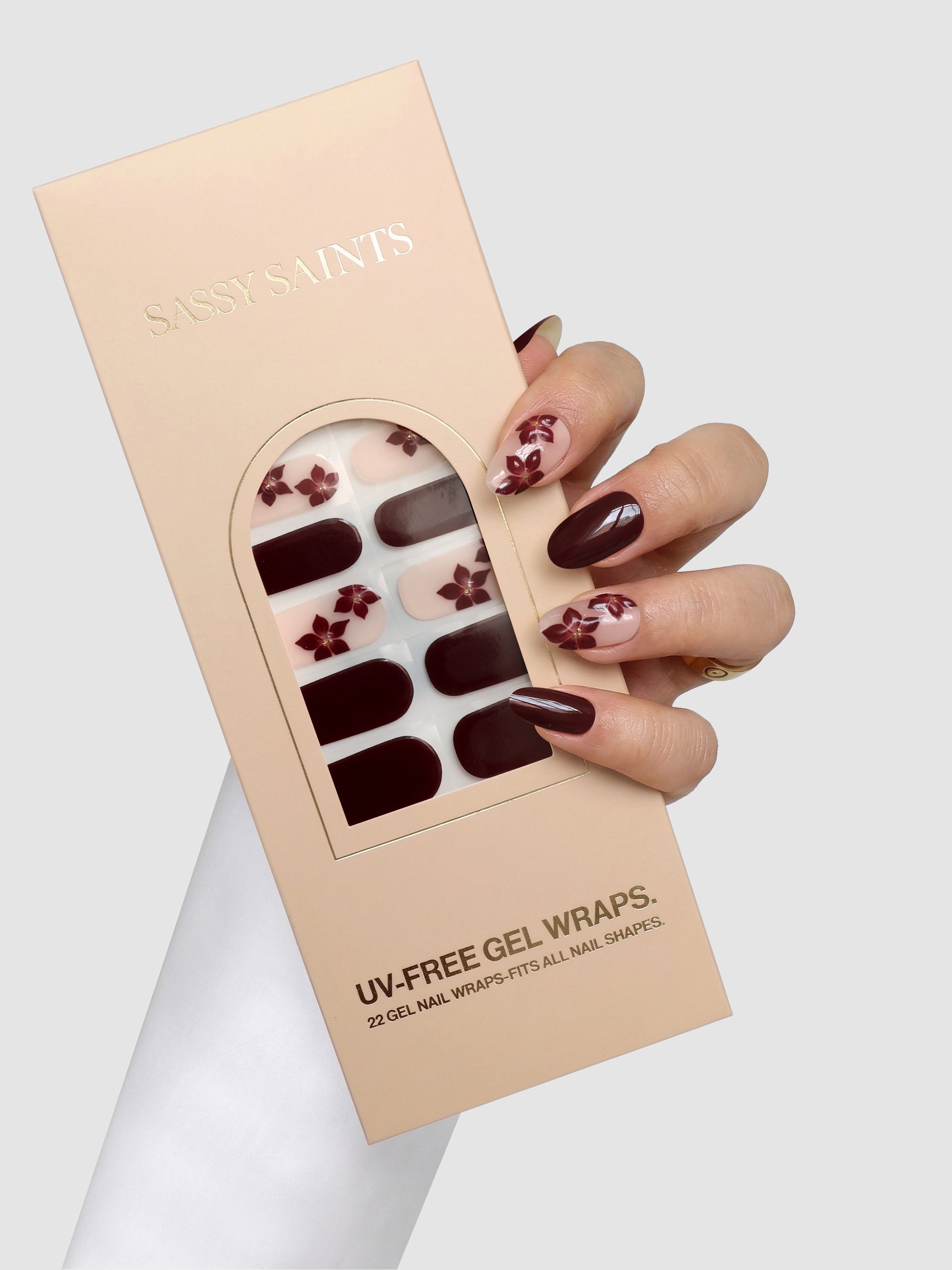 A hand with burgundy and floral nail art holds a beige box labeled “Sassy Saints – Brings The Salon Home” Cabernet Corsage Gel Wrap, containing burgundy and floral gel nail wraps.