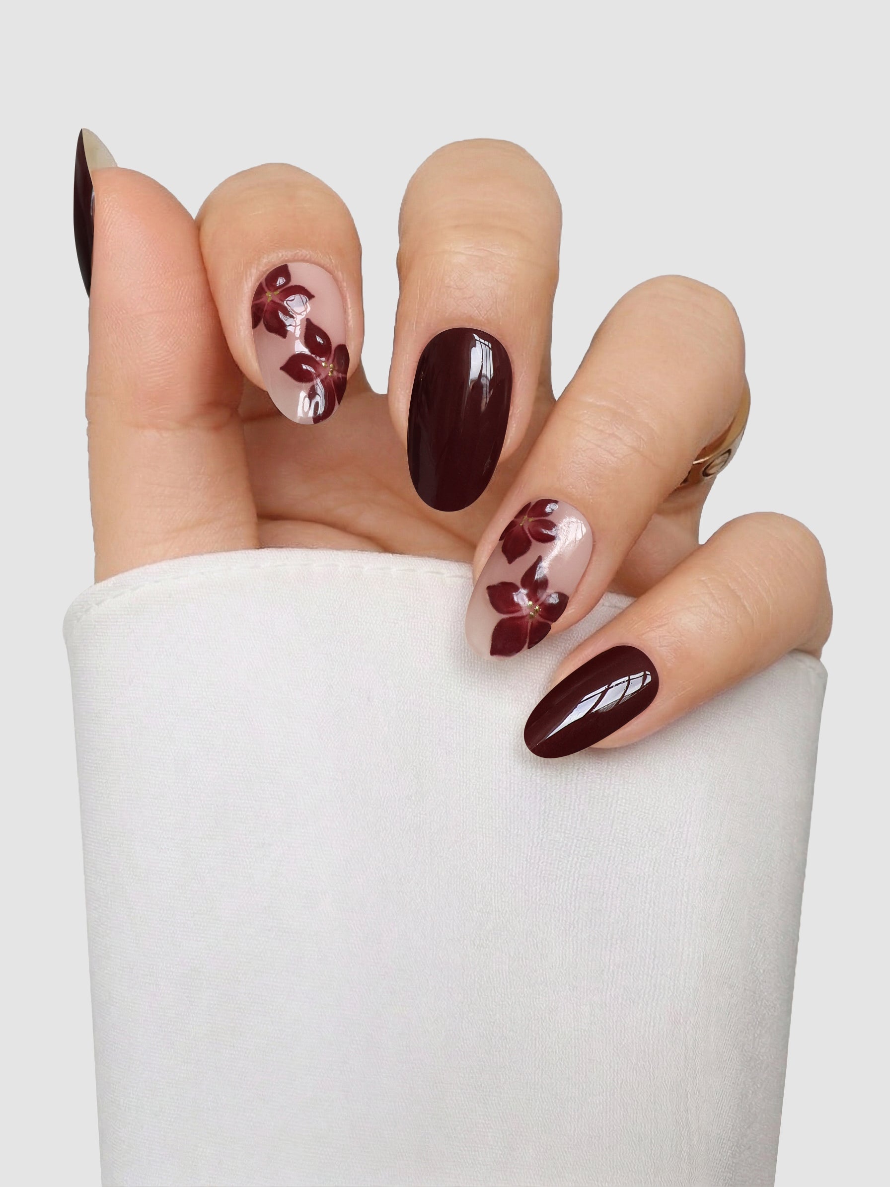 A hand with almond-shaped nails, featuring the Cabernet Corsage Gel Wrap by Sassy Saints - Brings The Salon Home—designed in dark burgundy and nude shades with elegant burgundy floral accents—is displayed against a light background. The person is wearing a white long-sleeve top.
