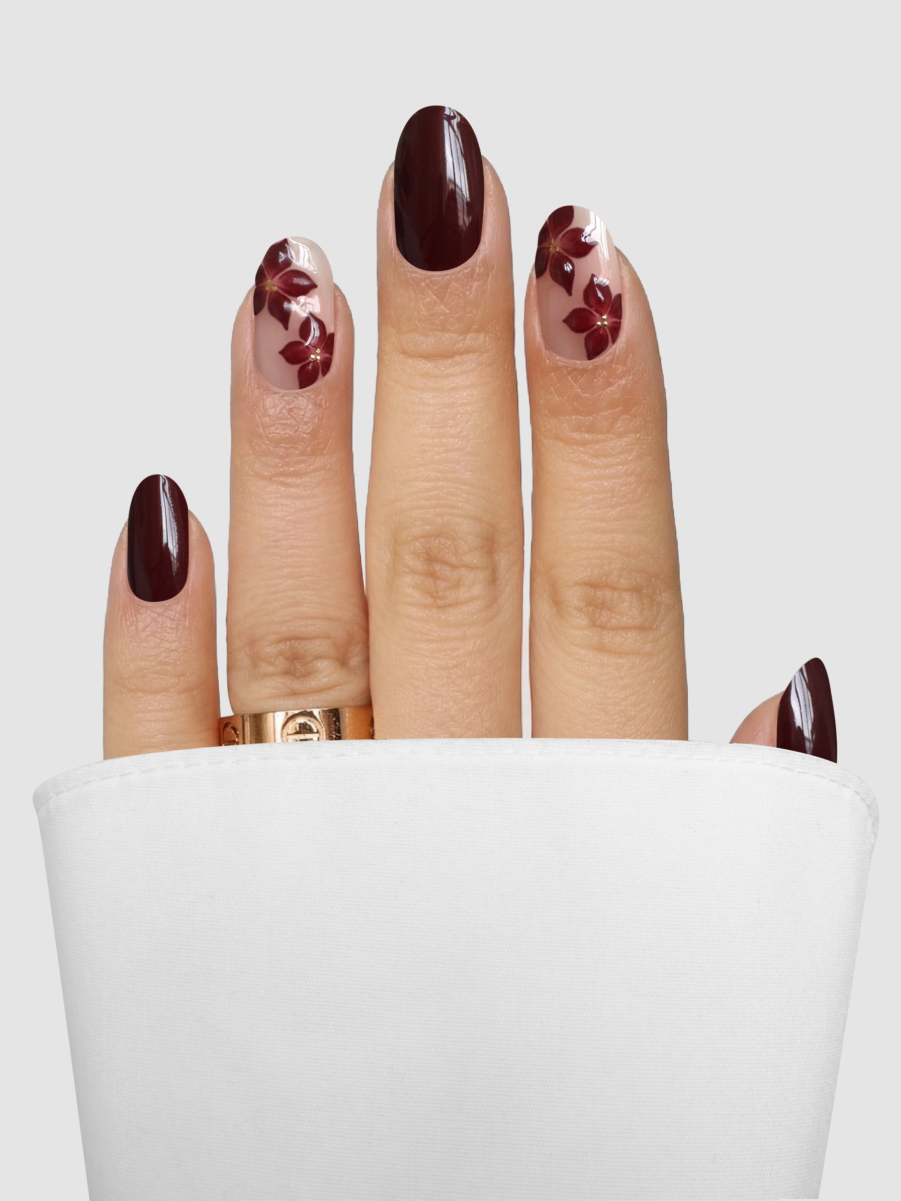 A hand with Cabernet Corsage Gel Wrap nail art from Sassy Saints - Brings The Salon Home rests on a white surface. Two fingers showcase floral designs on a clear base, while the others are finished in solid maroon. The hand is adorned with a gold ring.