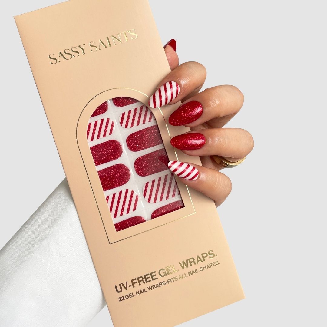 A hand with red and white striped and glittery red Candy Cane Gel Wraps holds a beige Sassy Saints – Brings The Salon Home box containing matching UV-free gel nail wraps.