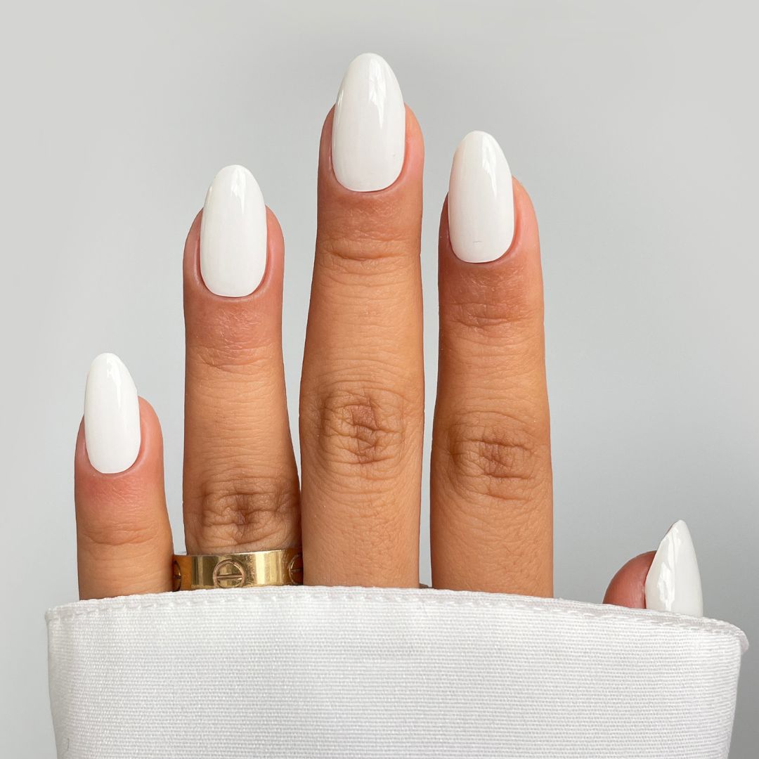 A hand with neatly manicured nails painted glossy white using Sassy Saints' Coconut Milk Gel Wrap is shown. The person wears a gold ring and a white sleeve, set against a plain, light background. Sassy Saints - Brings The Salon Home.