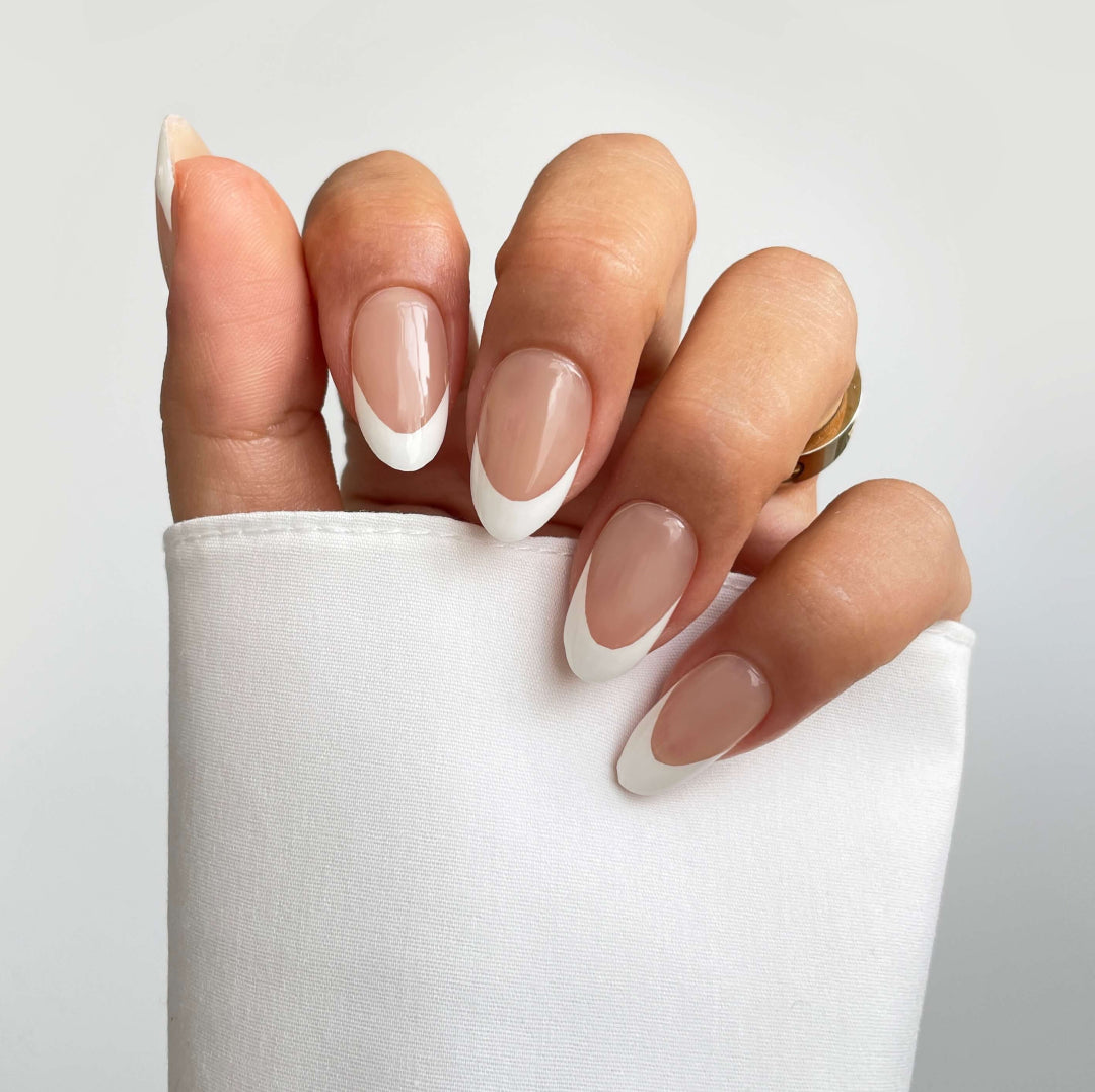 A hand showcases Sassy Saints - Brings The Salon Homes Classic French Gel Wrap nails with a natural pink base and white tips against a plain background. The person wears a white sleeve and a gold ring.